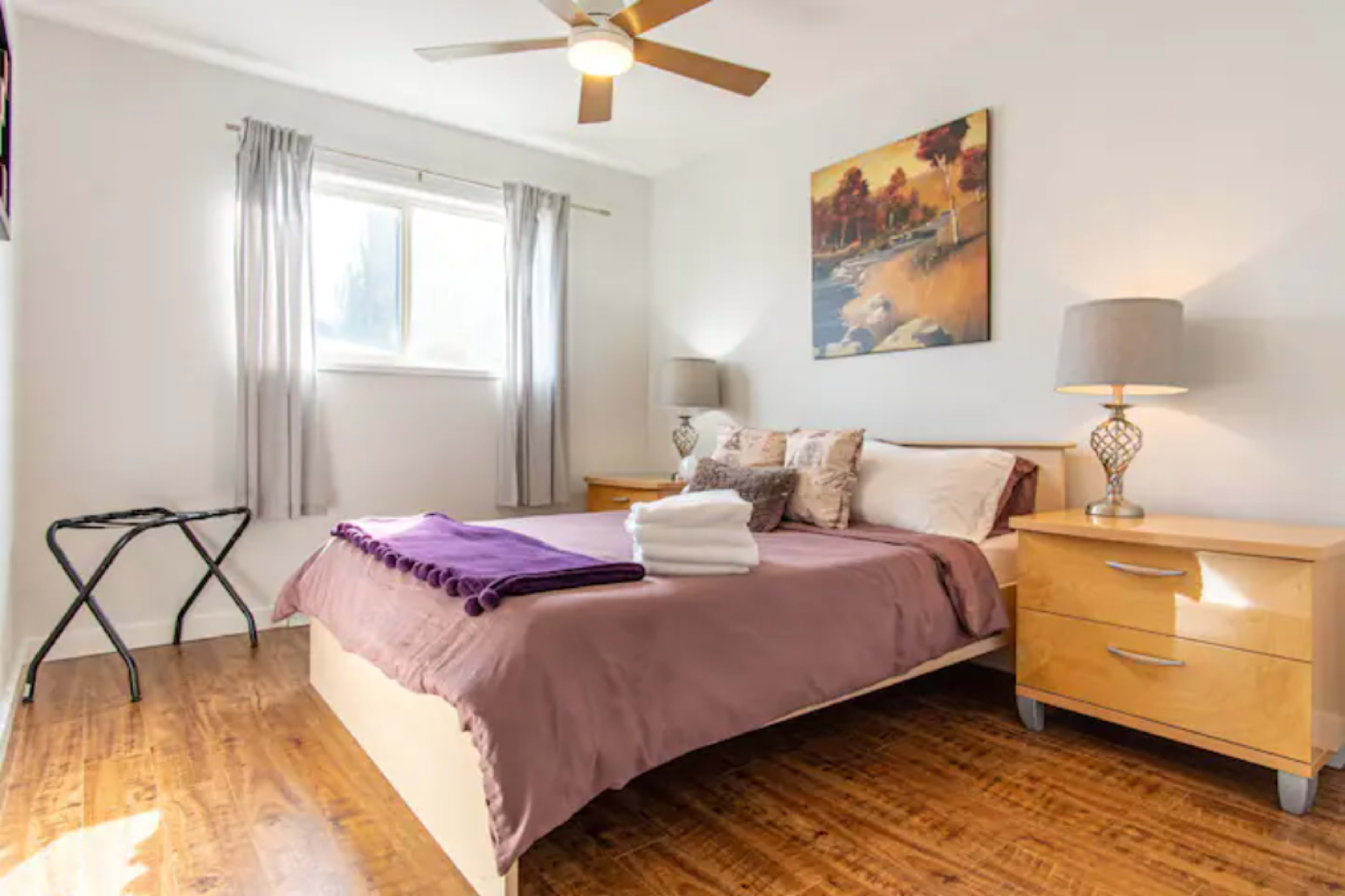A well-lit bedroom with a bed covered in a purple blanket, two nightstands with lamps, and a window with curtains.