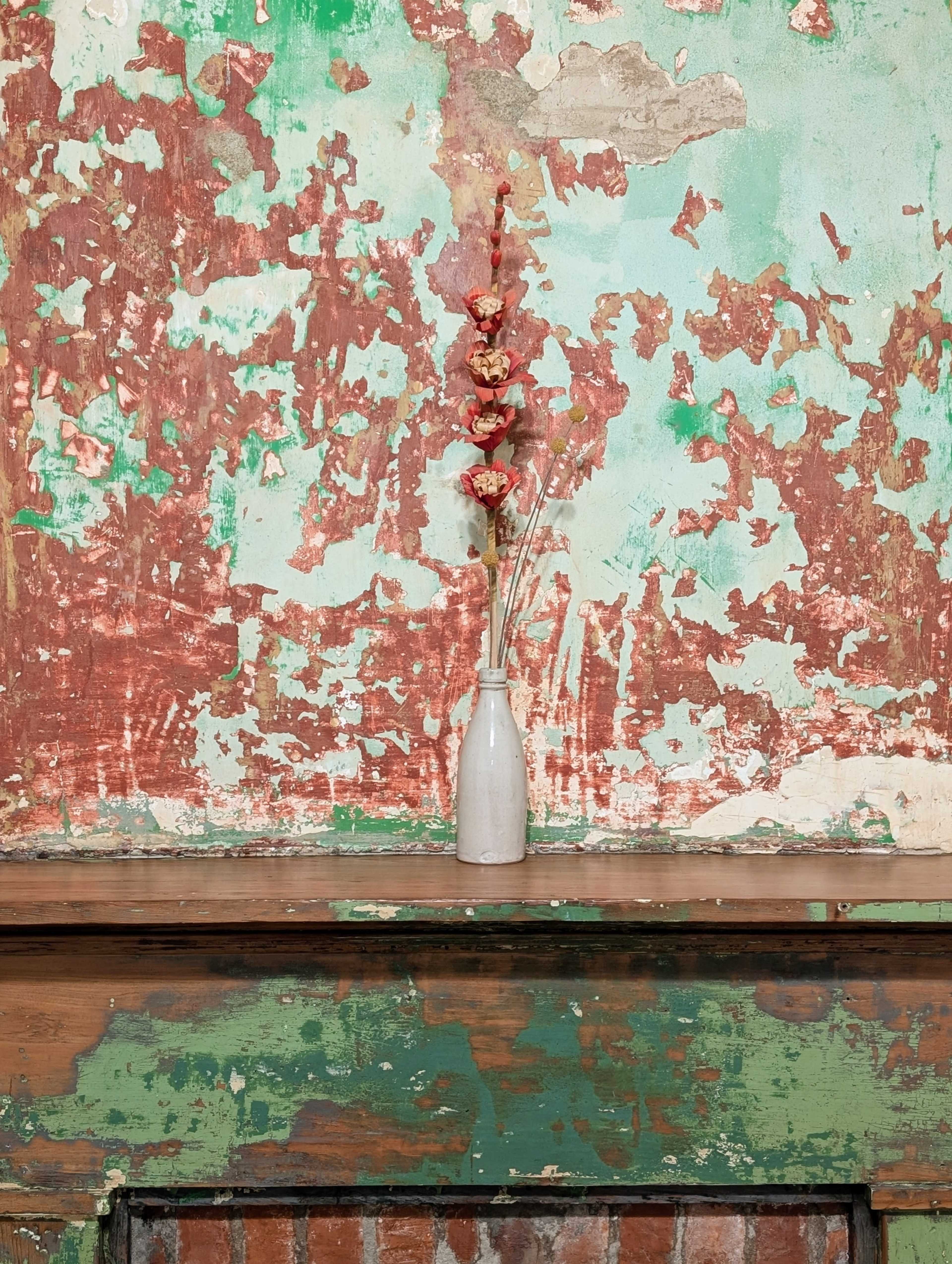 A simple white vase with dried flowers sits on a wooden ledge in front of a distressed, multicolored wall.
