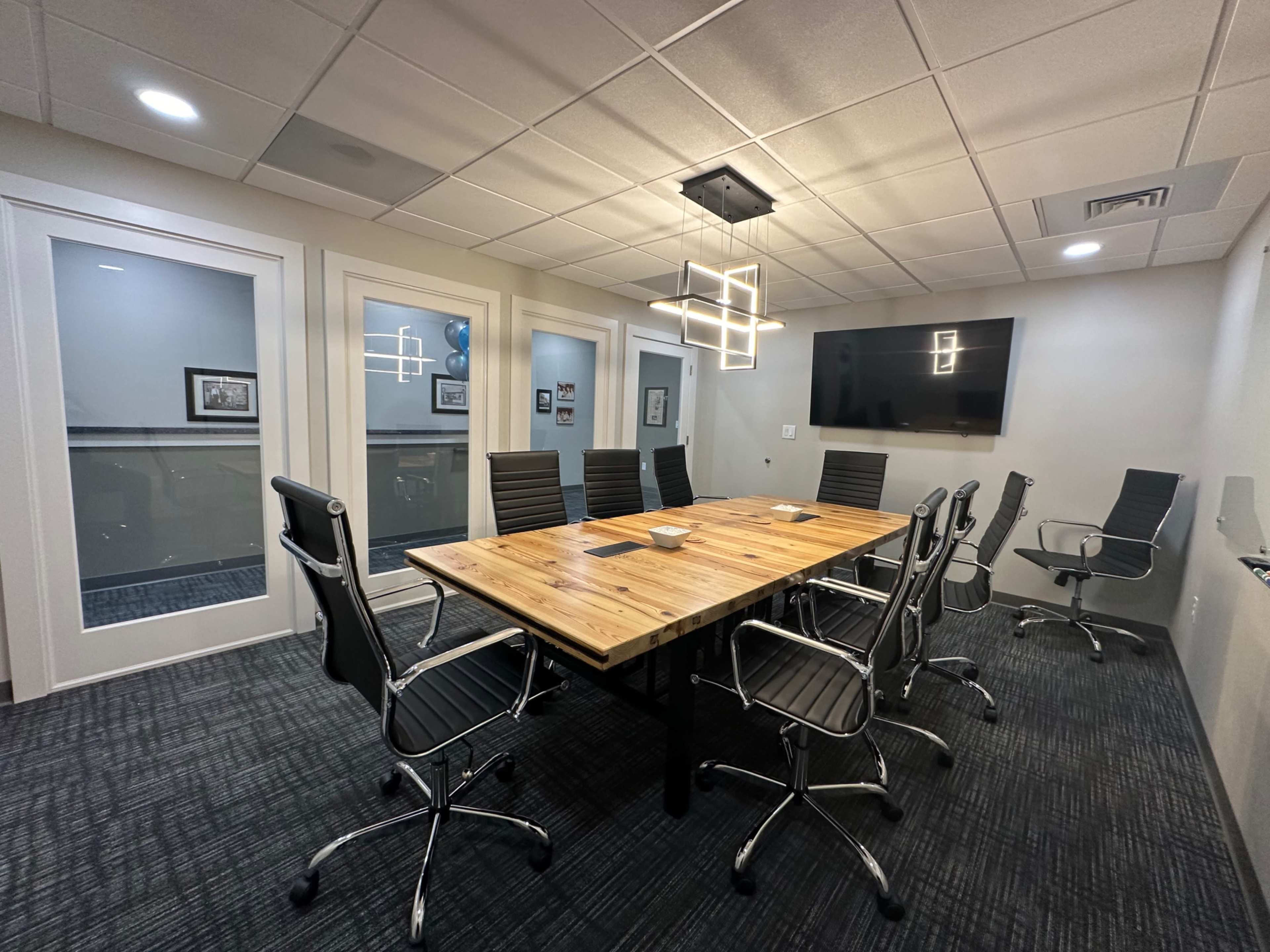 A conference room features a large wooden table surrounded by modern chairs, with glass walls, a television on one side, and decorative lighting.