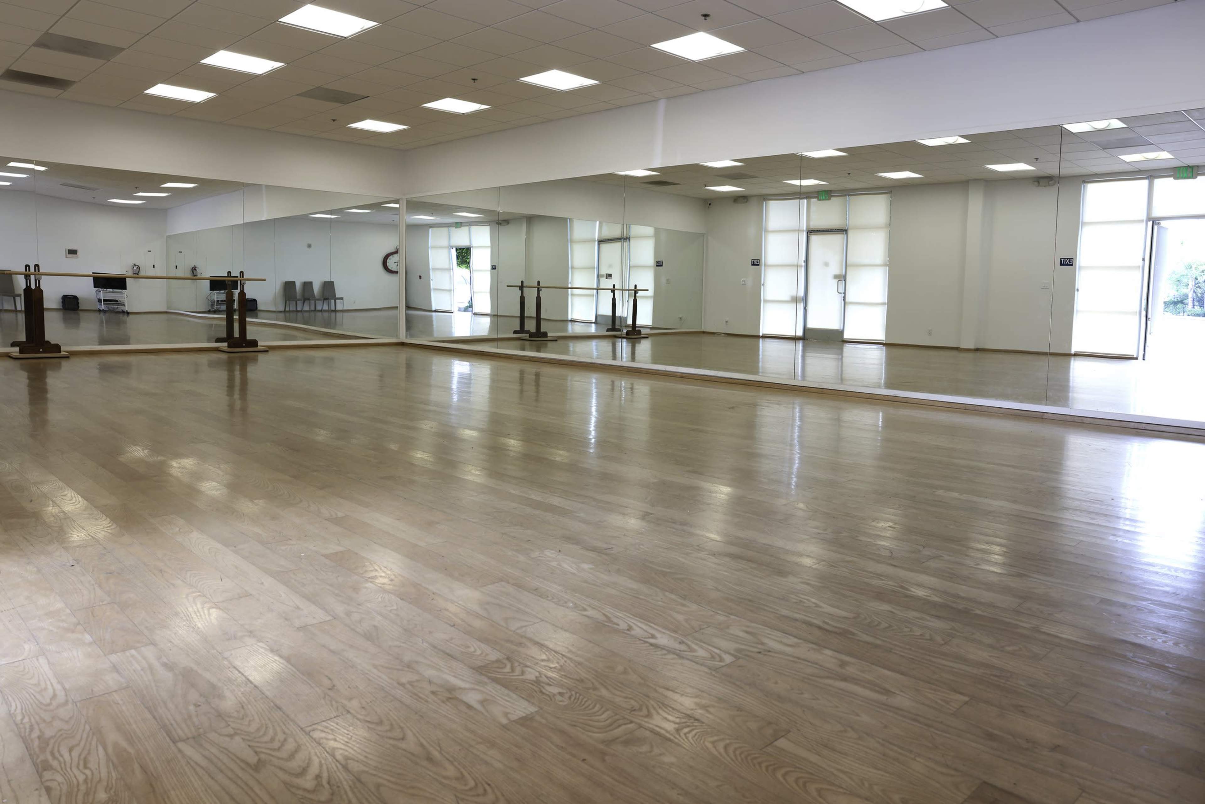 A spacious dance studio with wooden floors, large mirrors on one wall, and doors leading to an outside area.