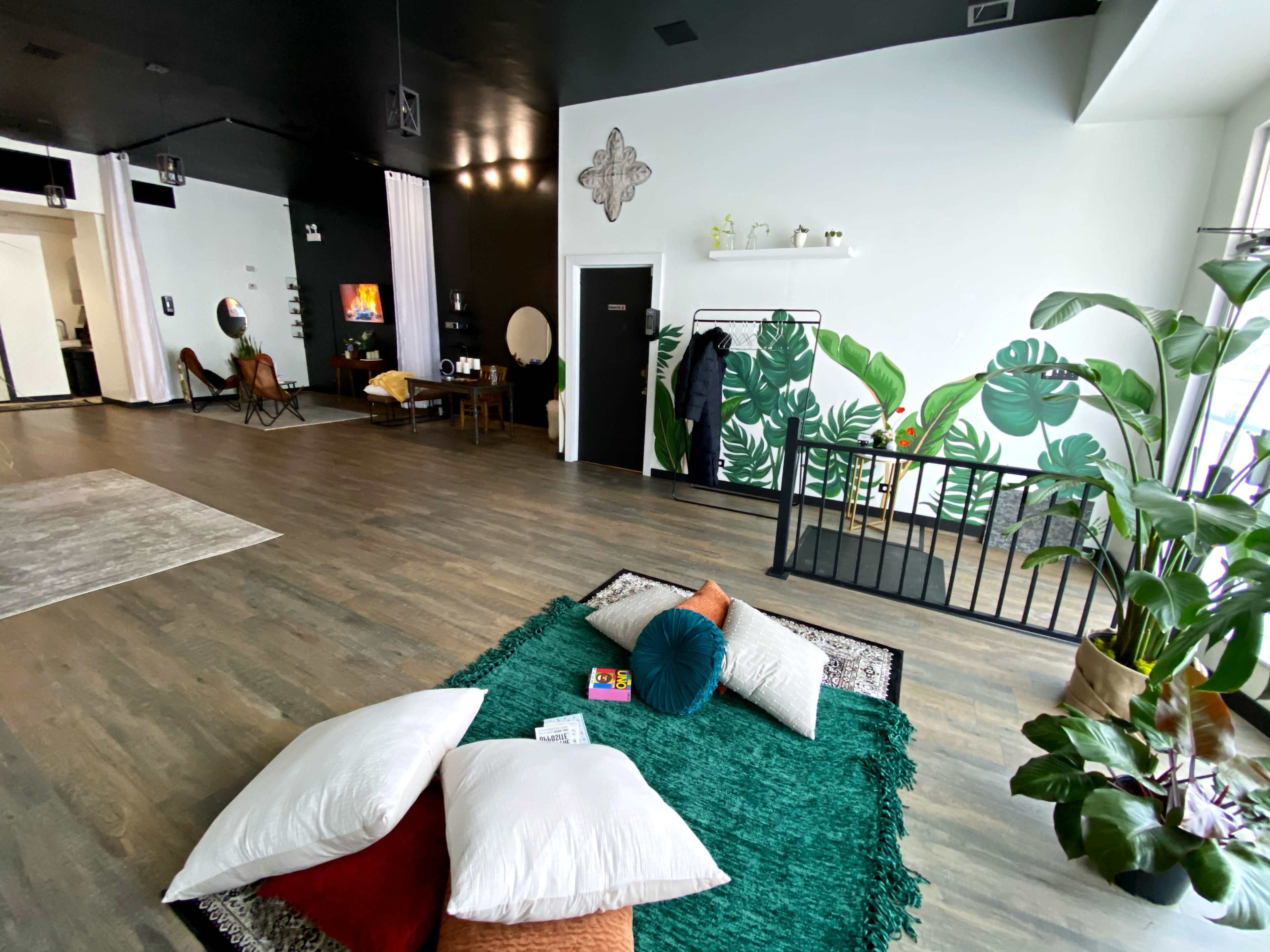 A spacious interior with a green rug and scattered cushions, accented by large plants and a wall featuring a tropical mural.