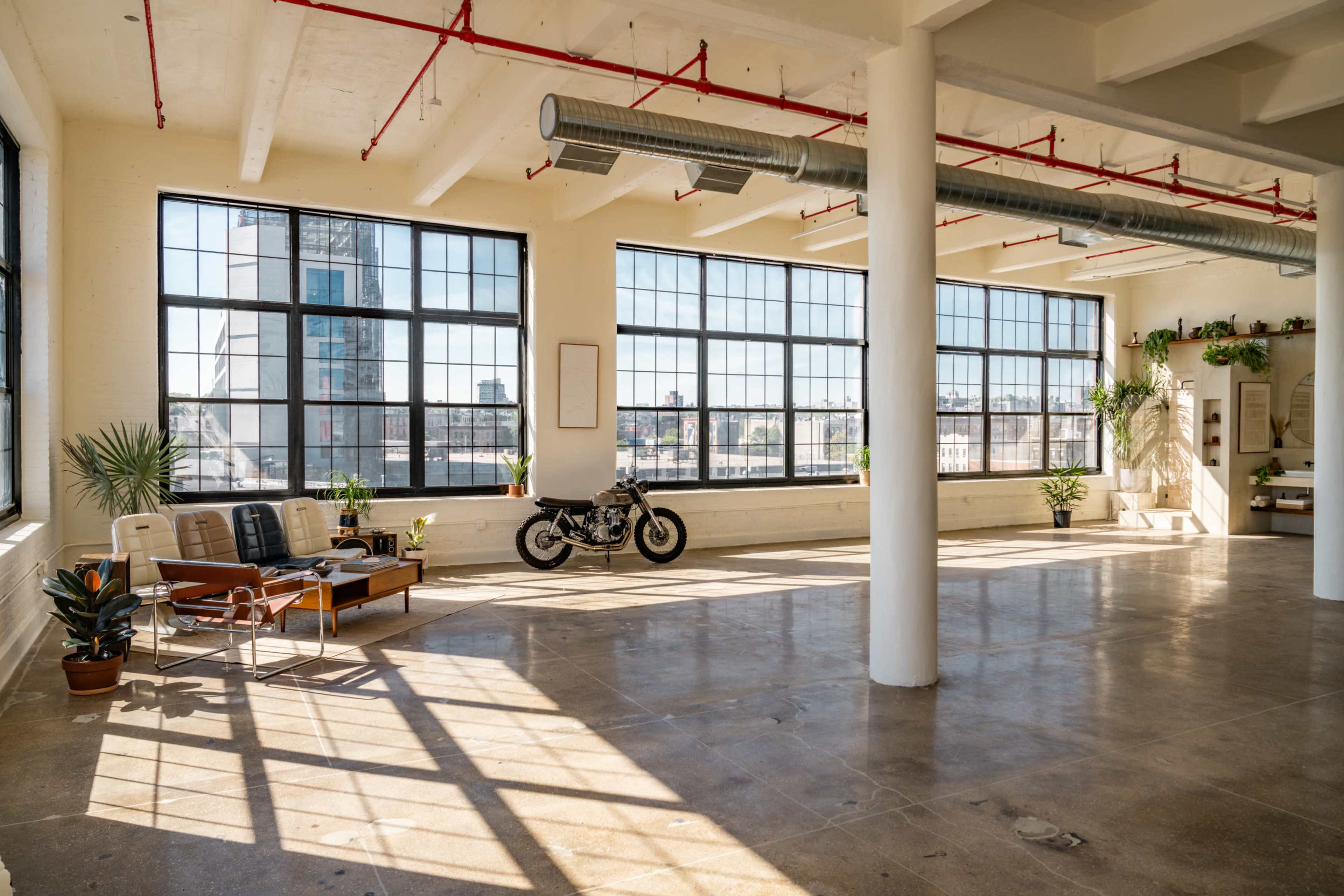 A spacious industrial loft features large windows, a motorcycle, and plants, with sunlight illuminating the concrete floor and minimalistic furniture.