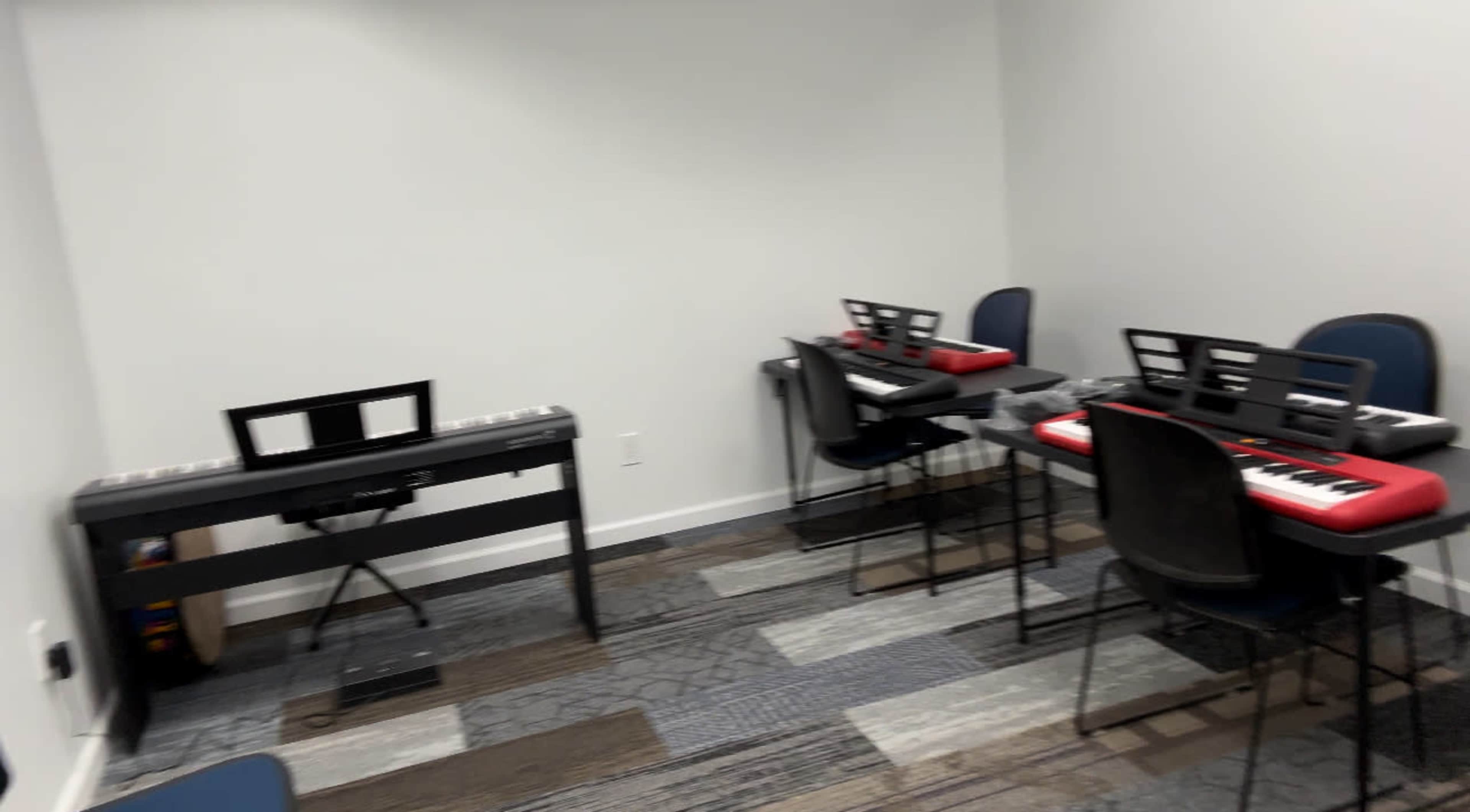A music room with two electronic keyboards on red stands and several chairs arranged around them.