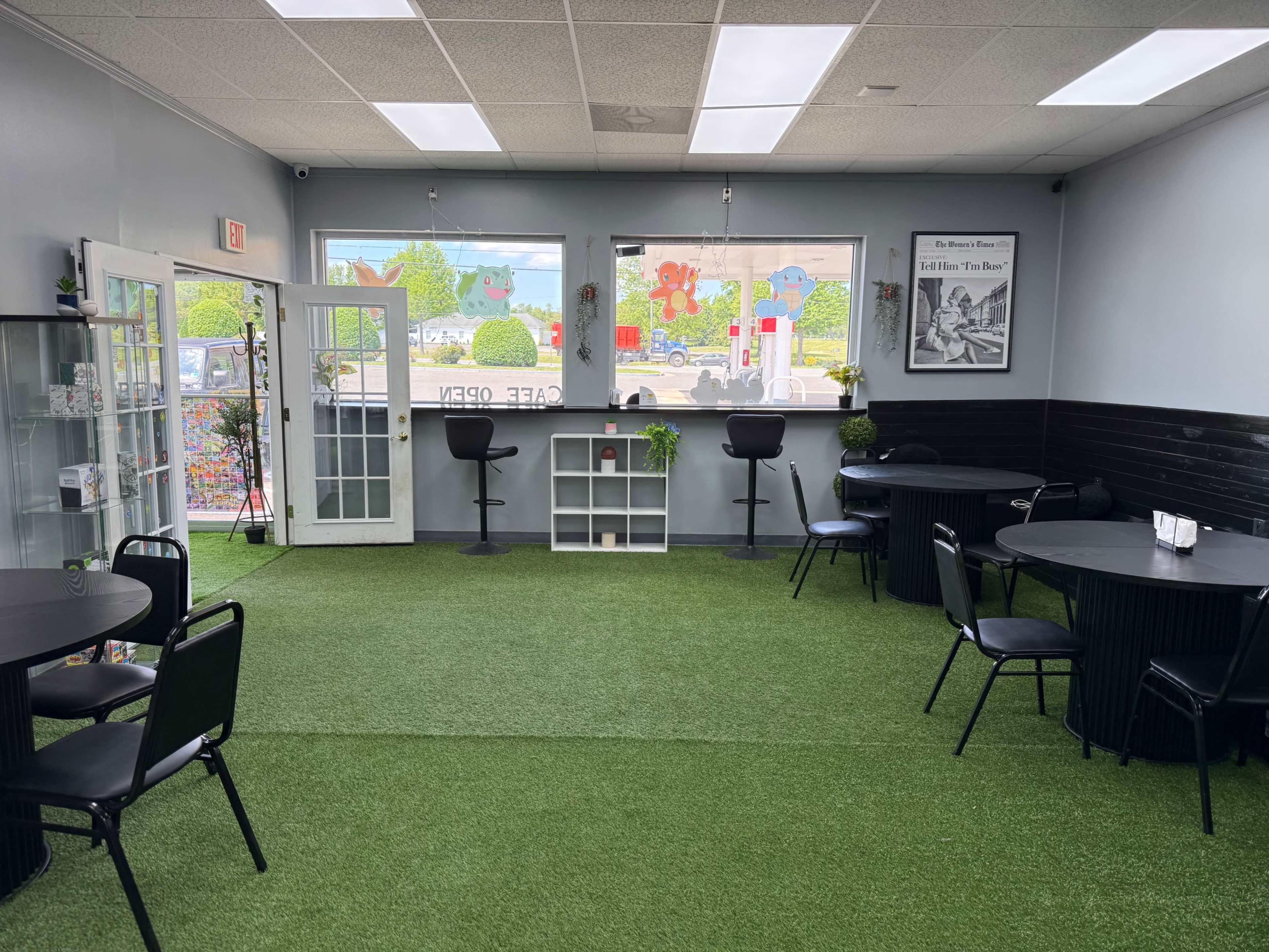 The image shows a small café with artificial grass flooring, several round black tables and chairs, and large windows displaying greenery outside.