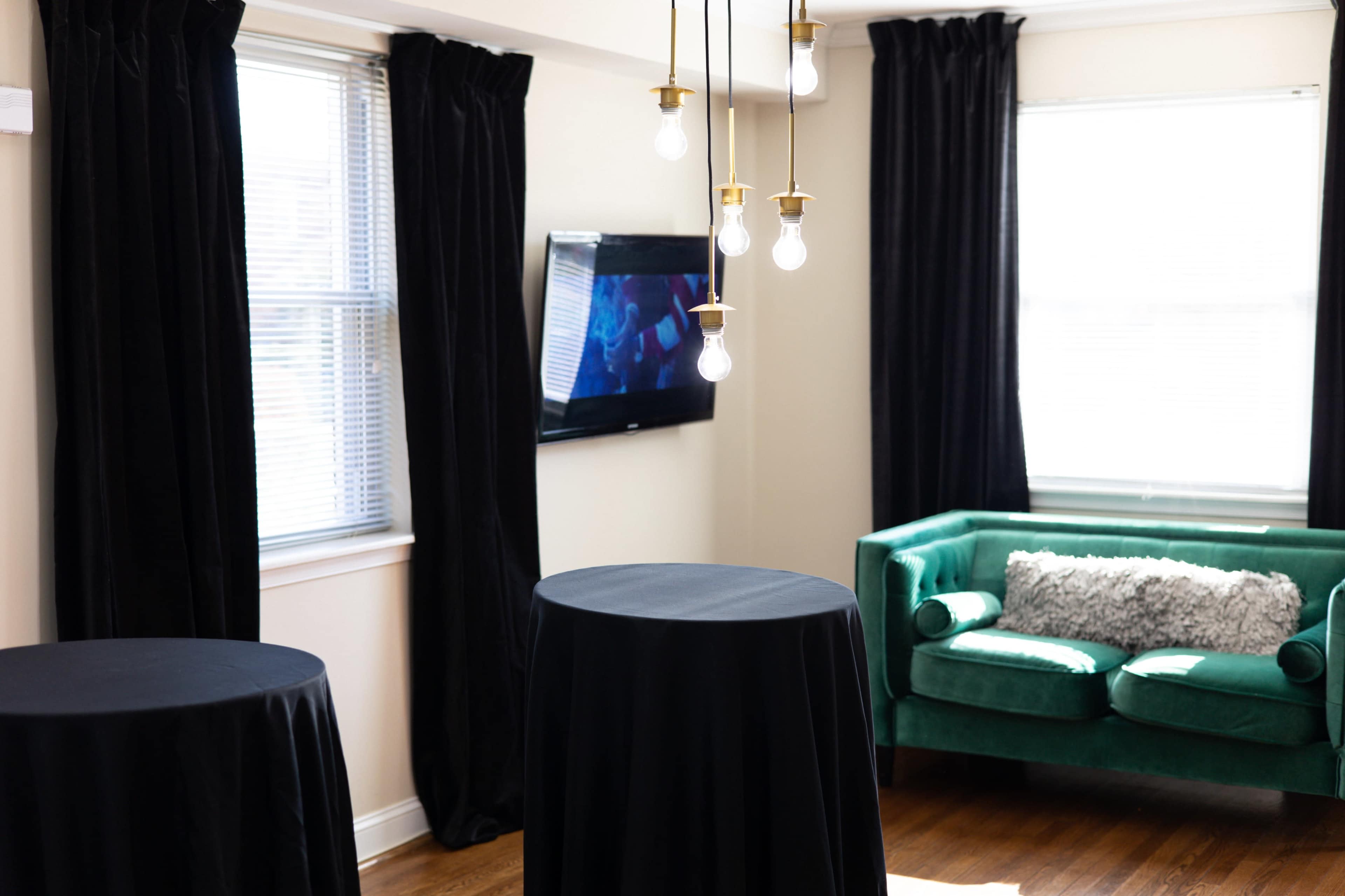 The image shows a well-lit room featuring two tall black tables, a green sofa, and a television mounted on the wall.