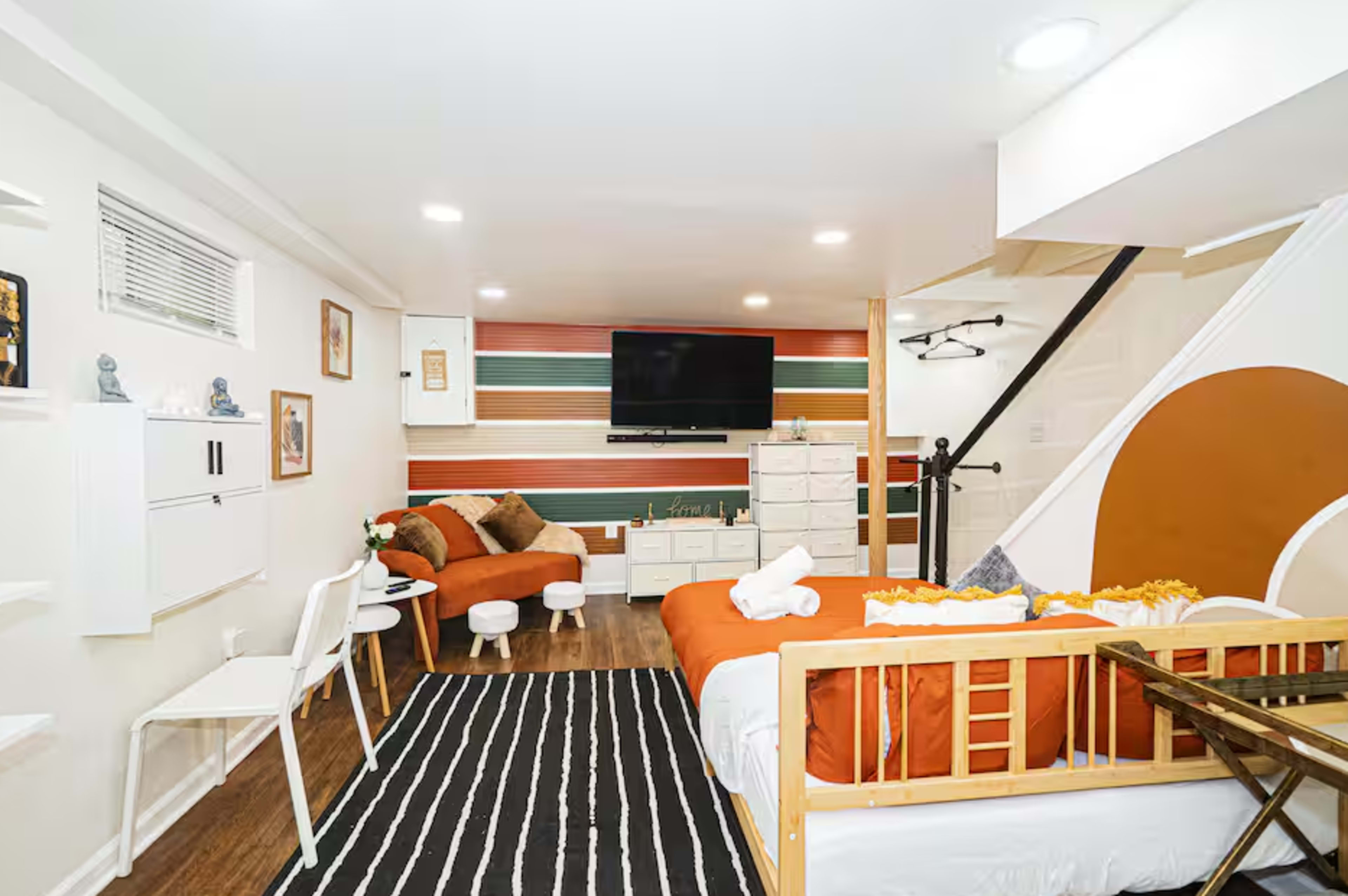 The image shows a modern basement living space with a sectional sofa, a TV mounted on the wall, decorative shelves, and a combination of a bed and a crib.