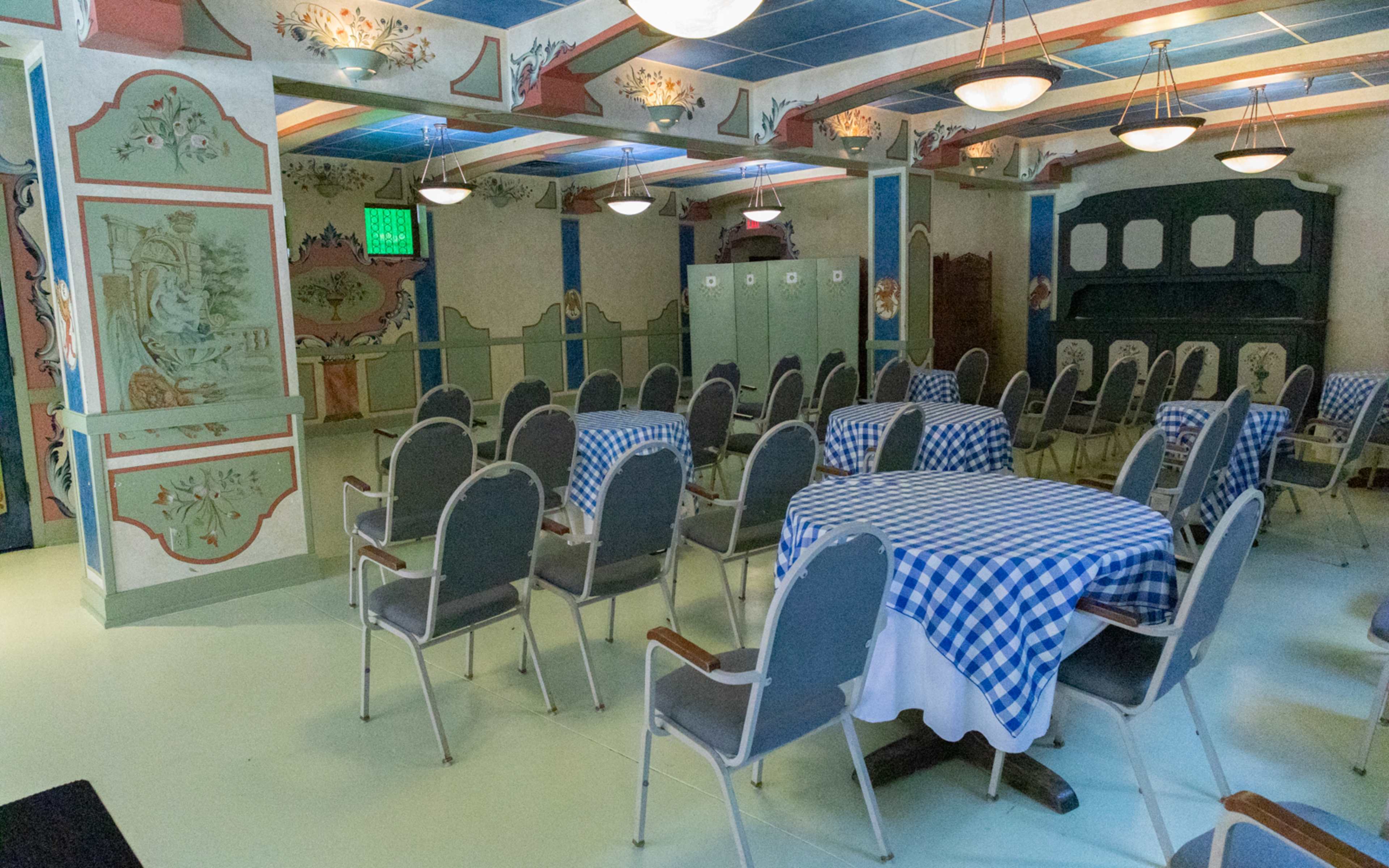A room with several round tables covered in blue and white checkered tablecloths, surrounded by chairs, with decorative walls and lockers in the background.