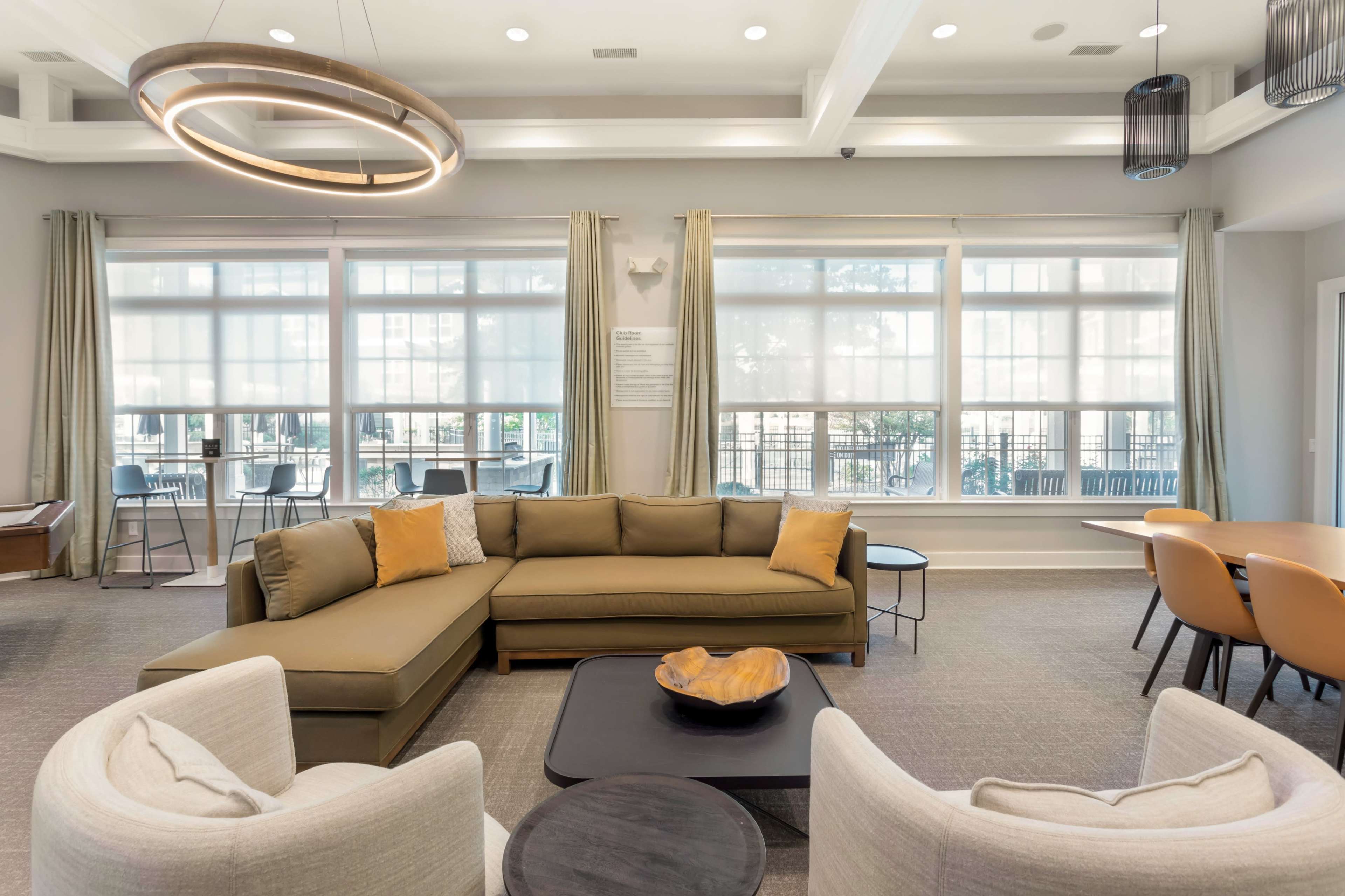 A modern communal lounge area featuring a sectional sofa, a round coffee table, and dining chairs beside large windows with natural light.