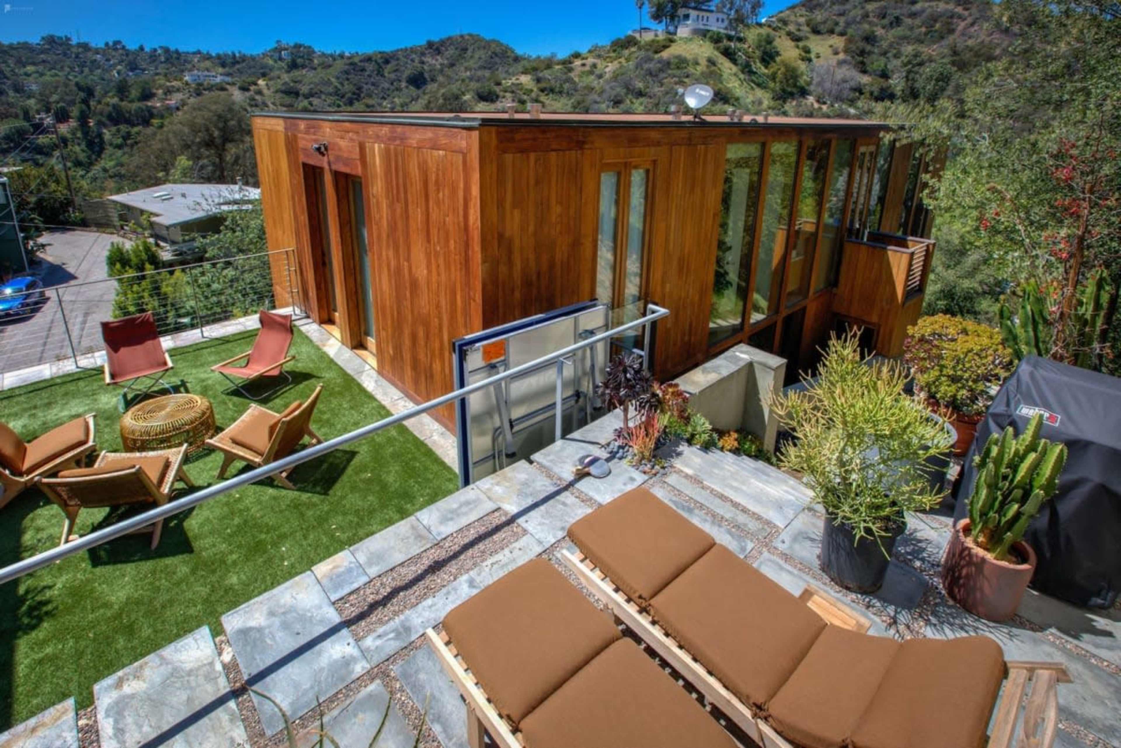 A modern wooden cabin with large glass windows is situated on a hillside, surrounded by greenery and outdoor seating areas.