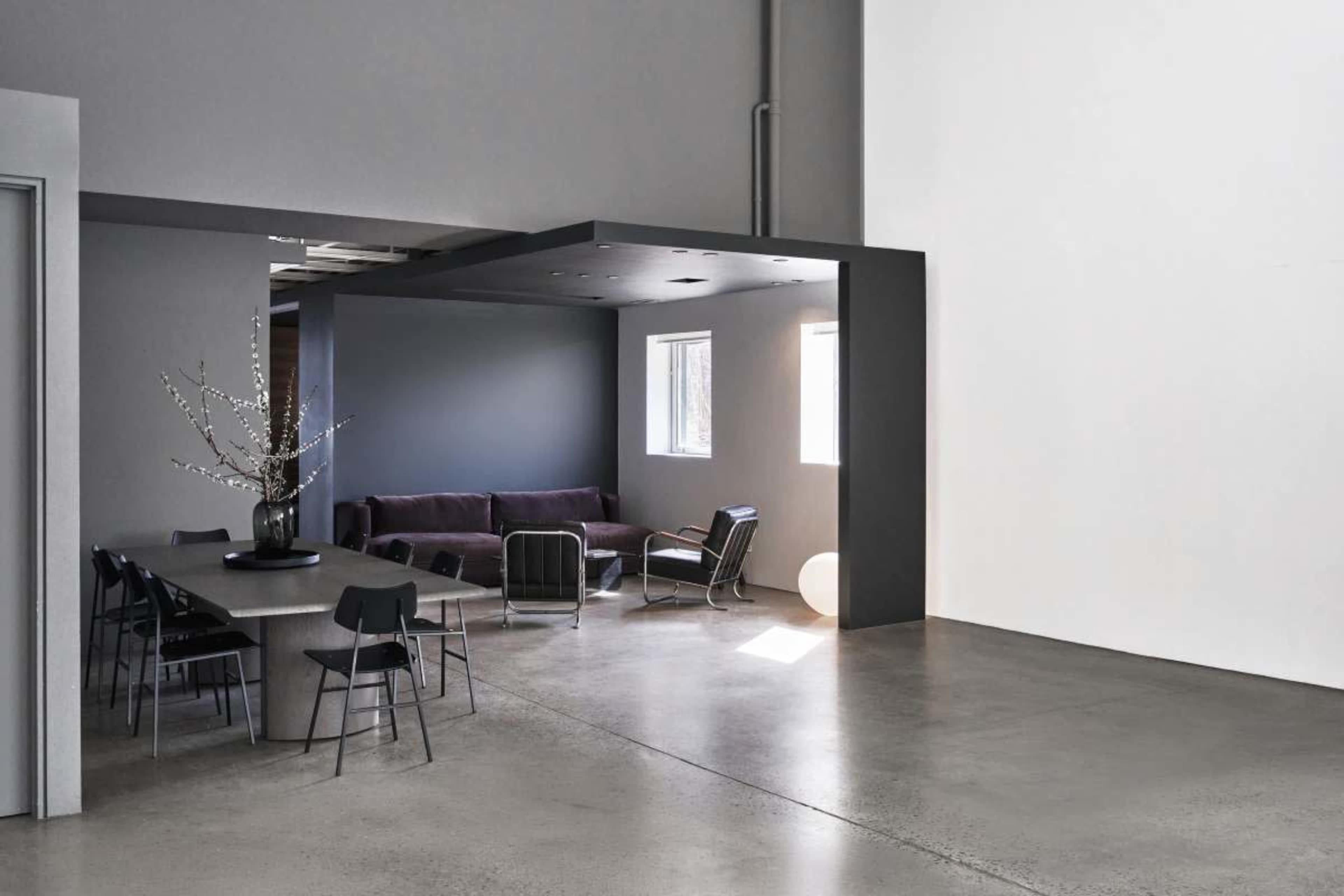The image shows a modern, minimalist interior featuring a dining area with a large table and black chairs, adjacent to a living space with a purple sofa and large windows.