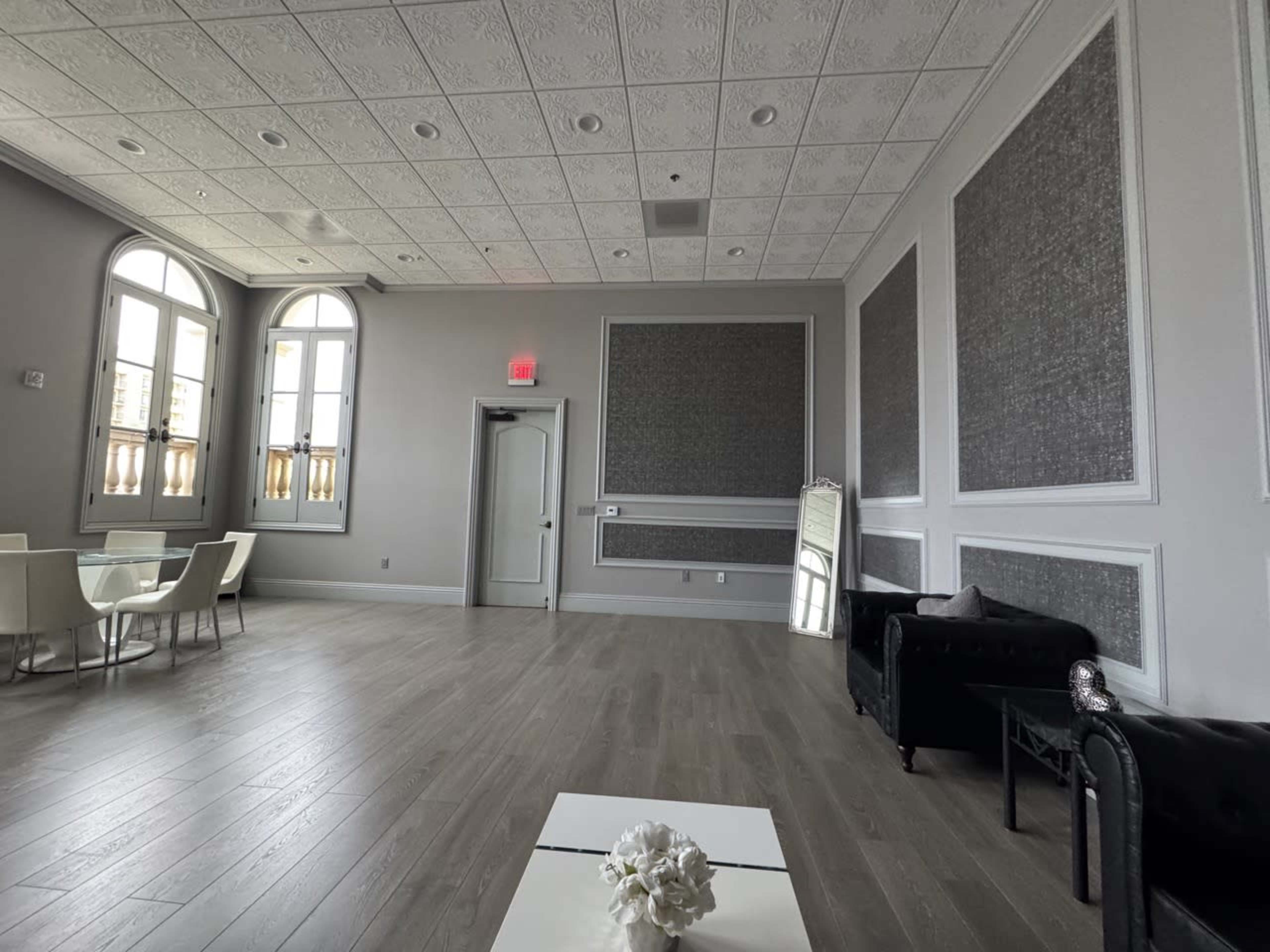 The image shows a spacious room with gray walls, light wooden flooring, and a table surrounded by white chairs, along with two black sofas and a large mirror.