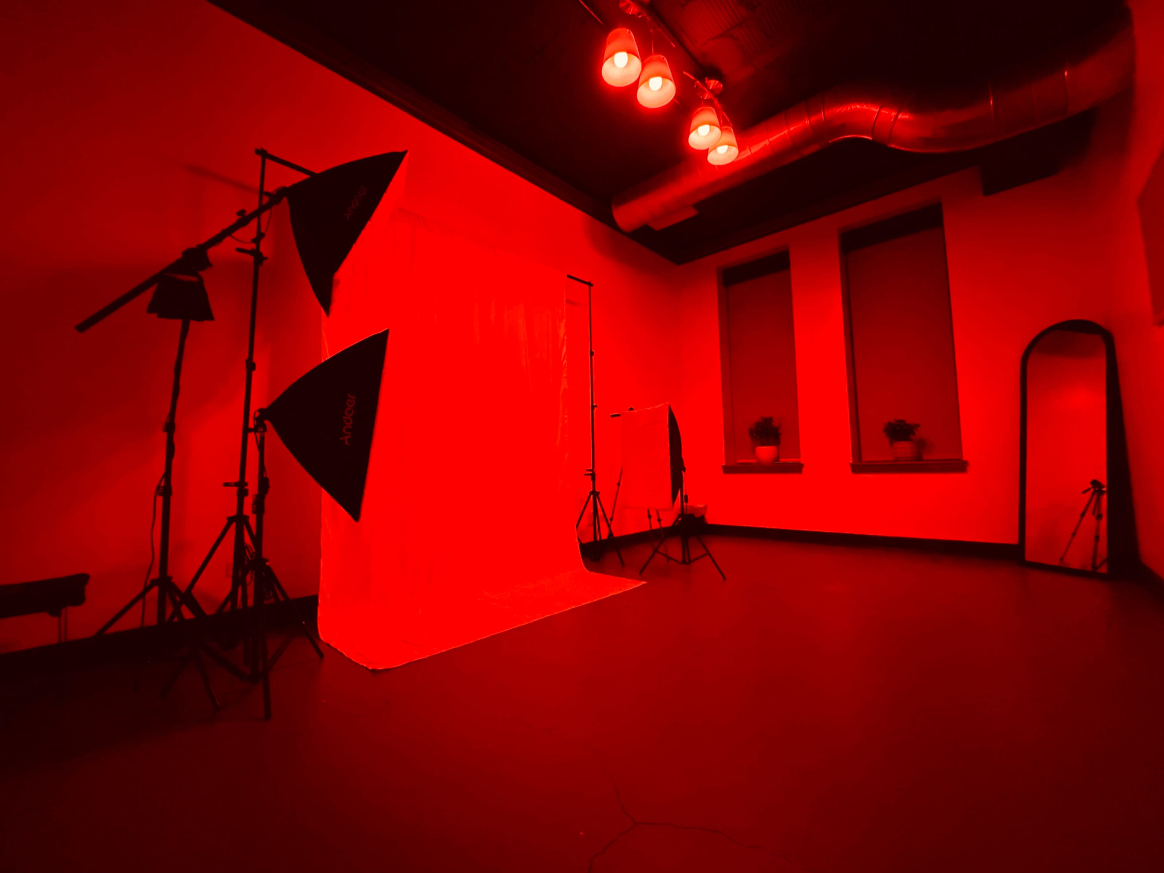 A photography studio with red lighting, featuring softbox lights, a white backdrop, and large windows framed by plants.