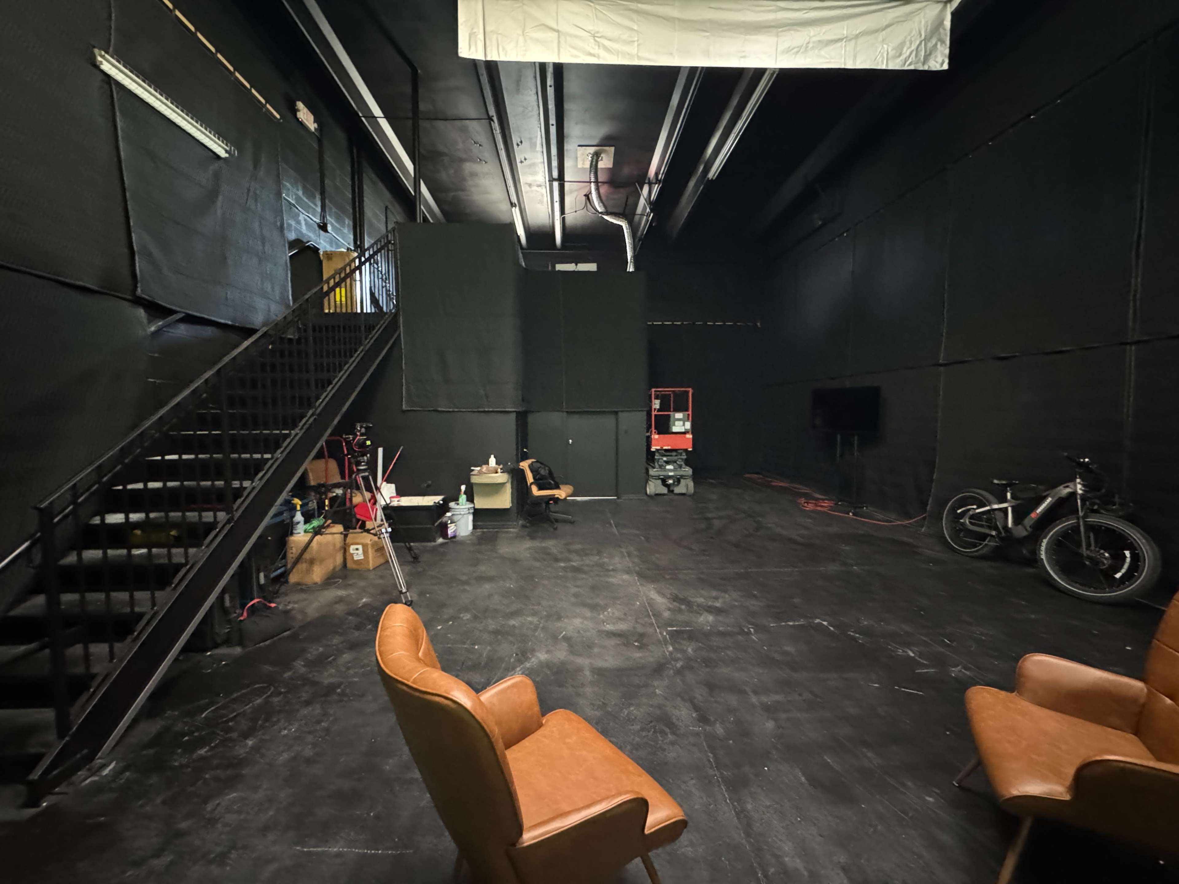 The image shows an empty, dimly lit studio space with black walls, a staircase on the left, two brown chairs, a bike, and various equipment along the sides.