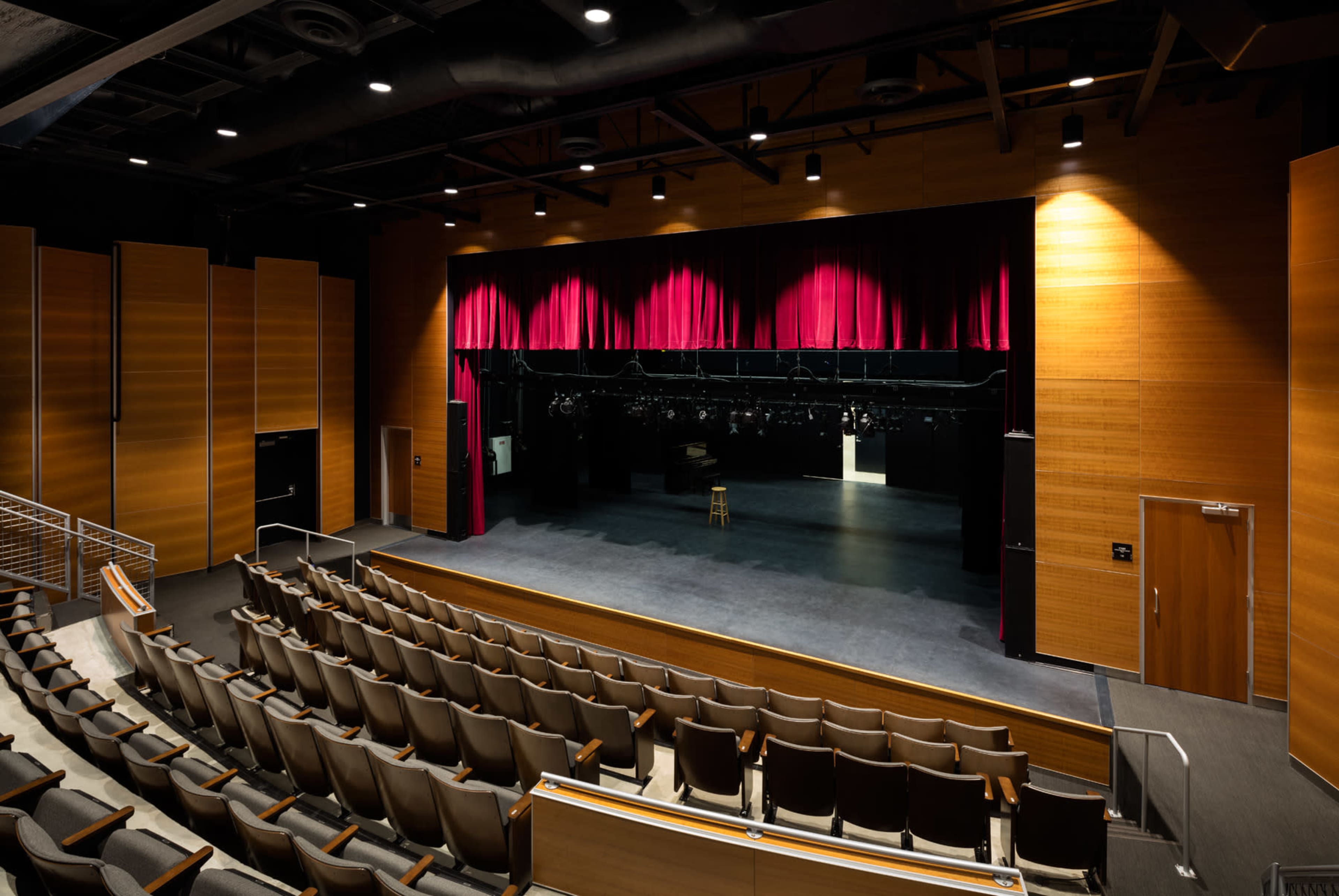 The image shows an empty theater with a stage featuring a red curtain and a single stool, surrounded by a seating area.