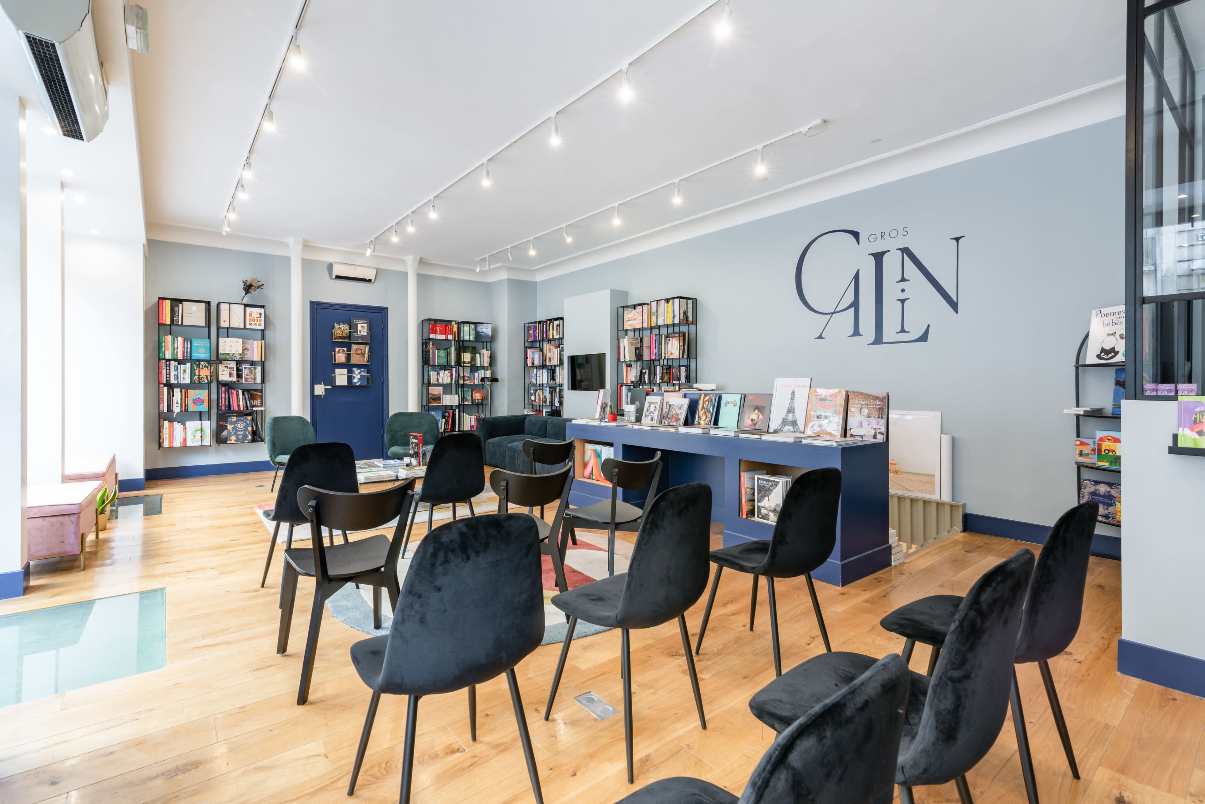 A well-lit room with rows of black chairs facing a display of bookshelves and tables containing books, set against a blue wall featuring the word "C A I N."