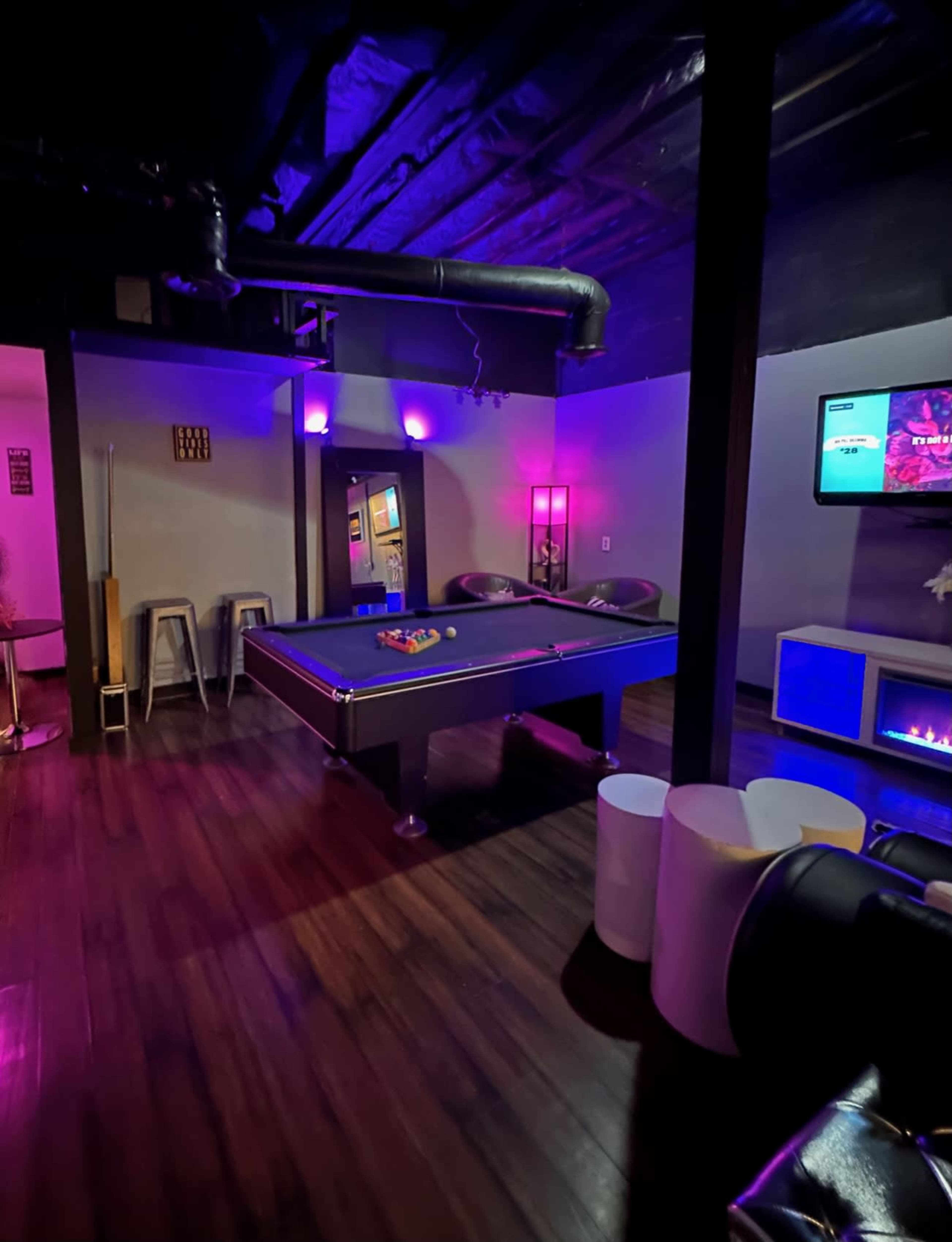 A recreation room featuring a pool table, bar stools, and a television, illuminated with purple lighting.