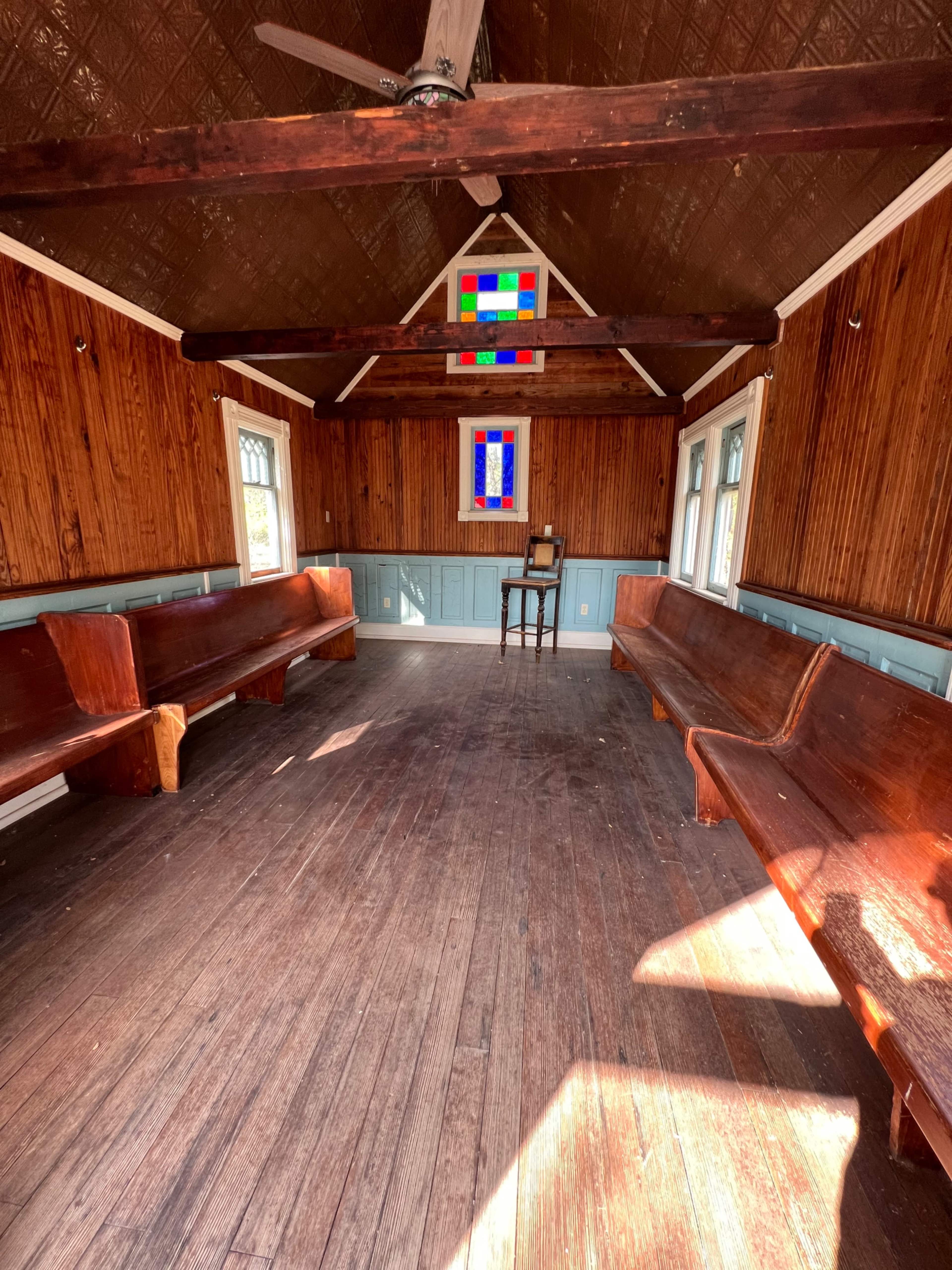 The image shows the interior of a wooden room with two long benches, a single chair, and a stained glass window.