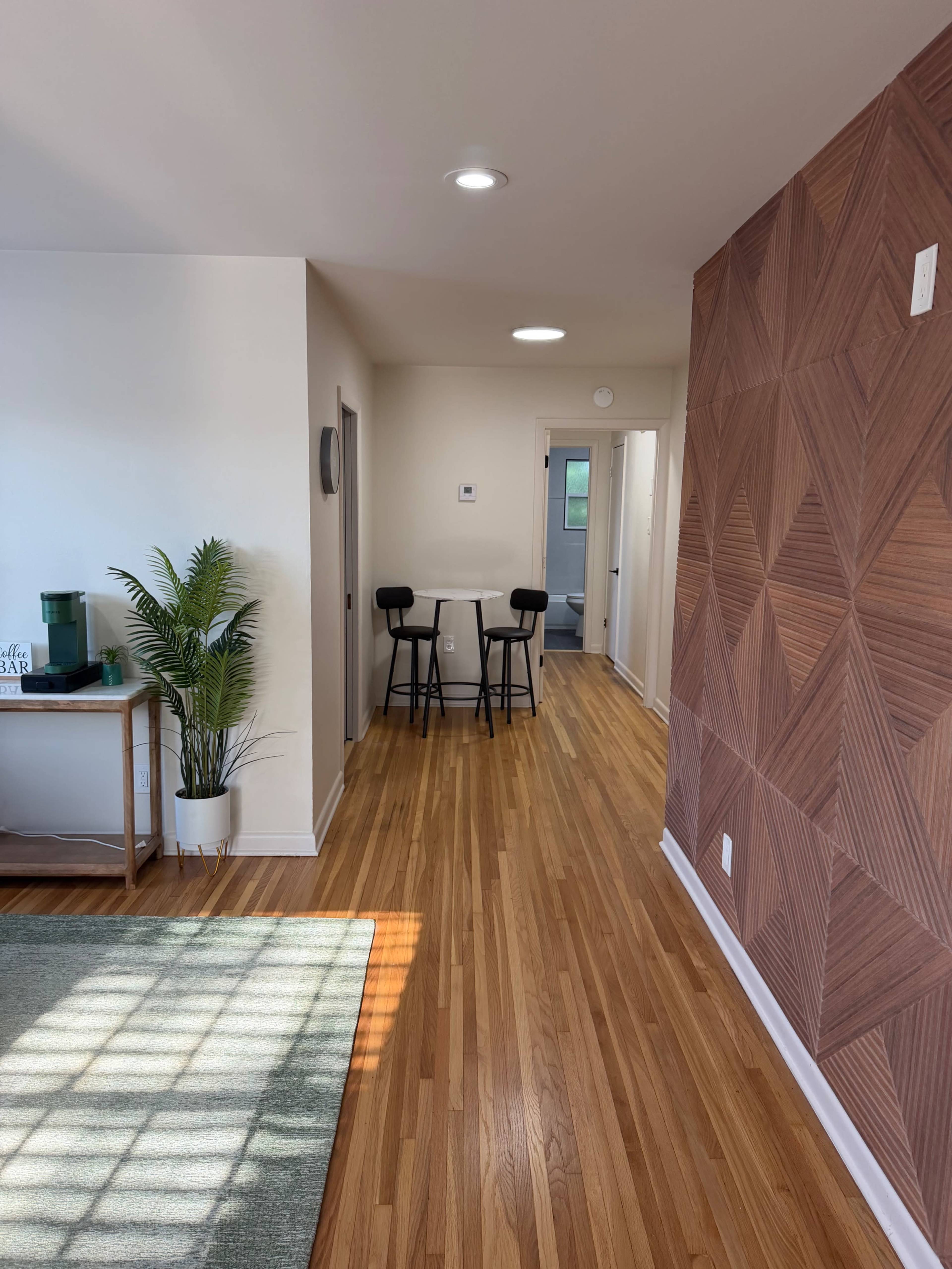 The image shows a well-lit interior space featuring a wooden floor, a patterned accent wall, and a small dining table with two black chairs in a hallway.
