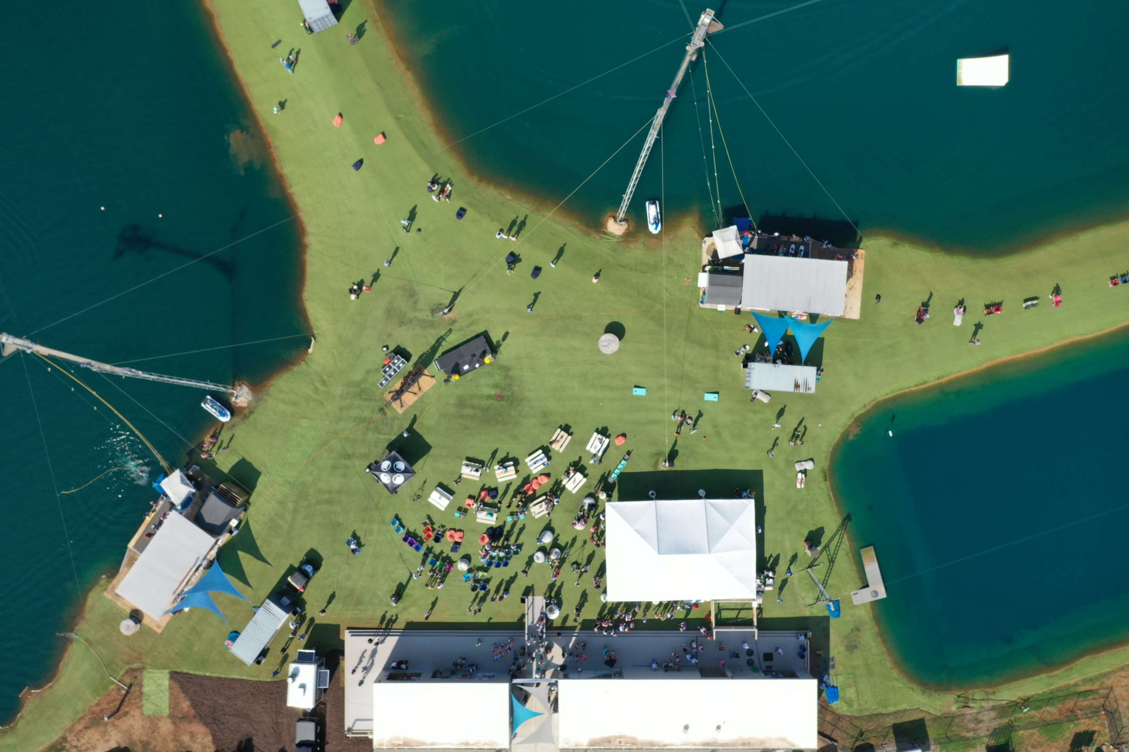 An aerial view of a water sports facility with people gathered on green grass and several water bodies surrounding the area.