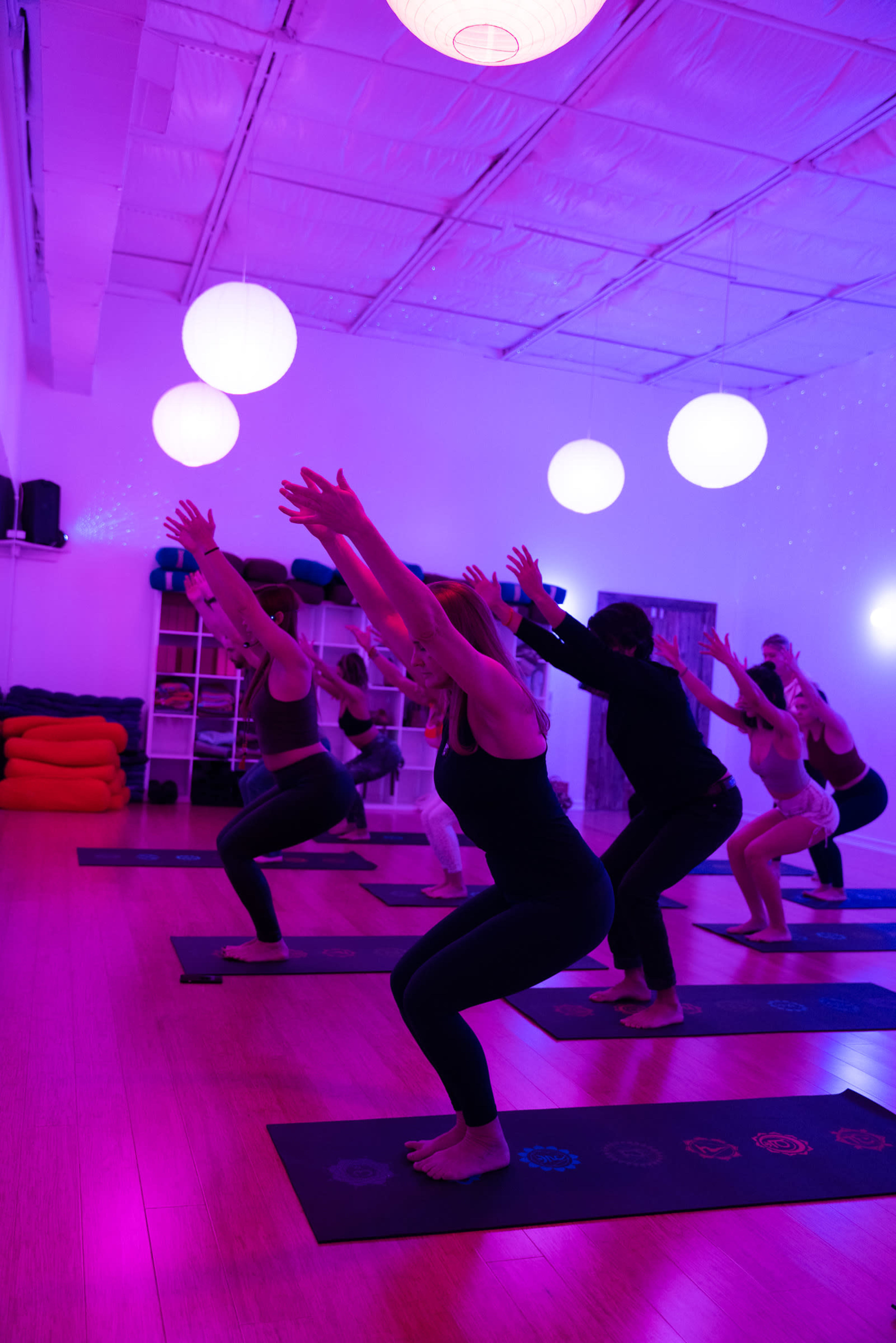 A group of individuals performs yoga poses in a studio lit with purple lighting and hanging round lamps.