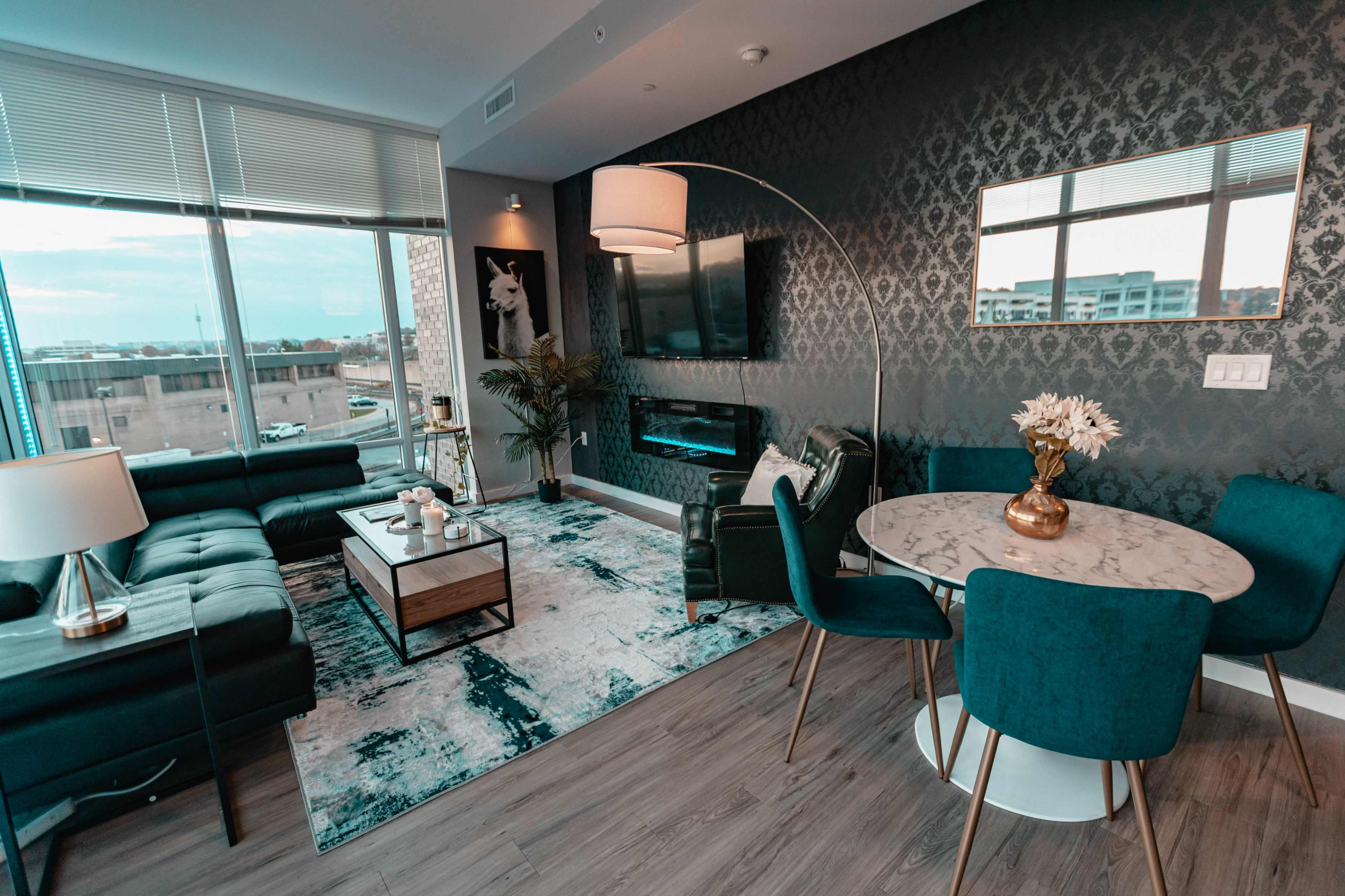 A modern living room featuring a green couch, a round dining table with teal chairs, a marble coffee table, a wall-mounted TV, and large windows that provide natural light.