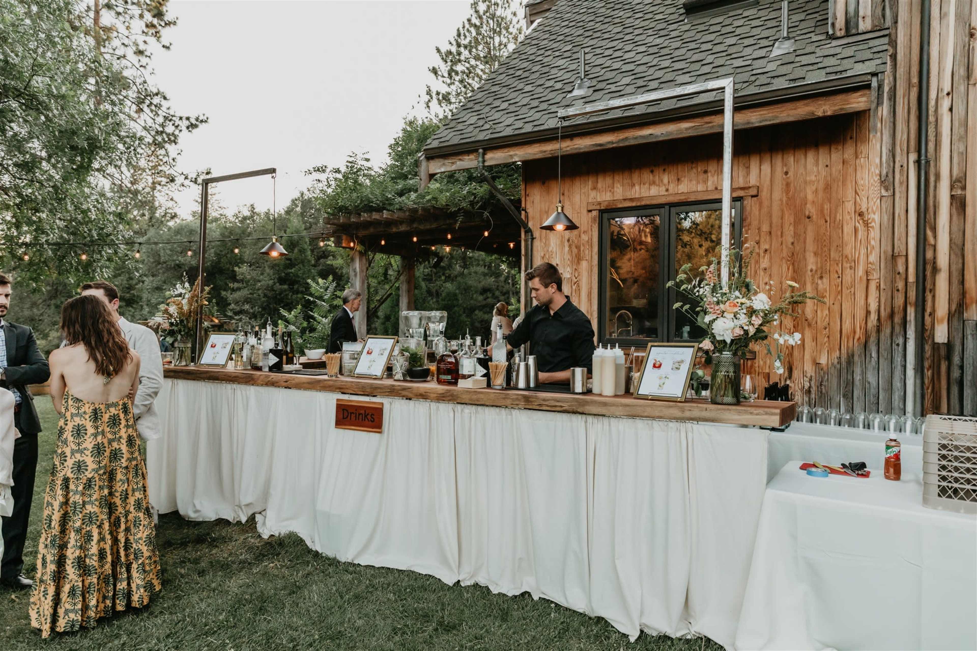 A wooden outdoor bar with a white tablecloth displays various drinks while guests mingle in front of a rustic house surrounded by greenery.