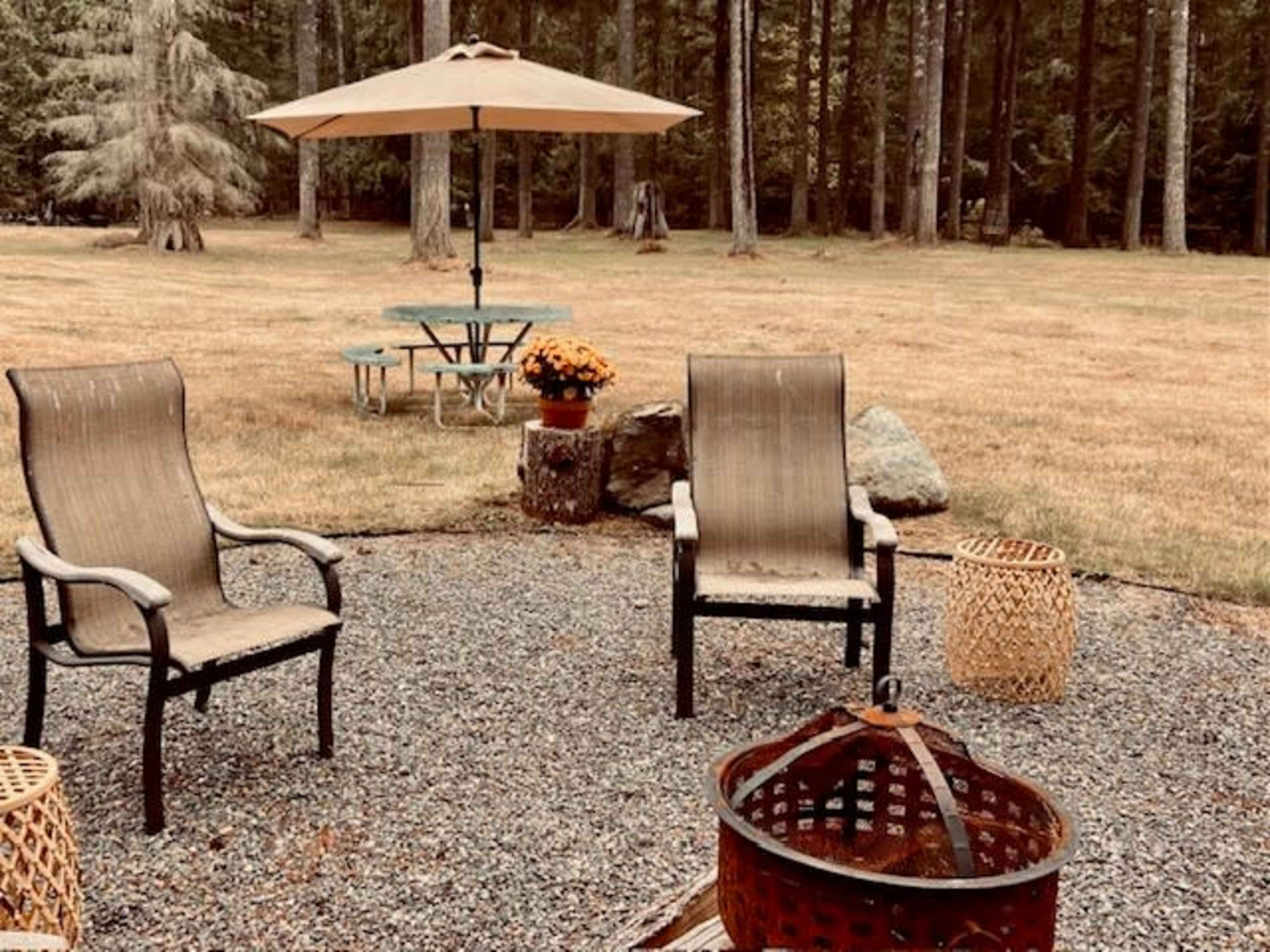 The image shows a gravel patio area with two chairs facing a fire pit, an umbrella for shade, and a picnic table in the background surrounded by trees.