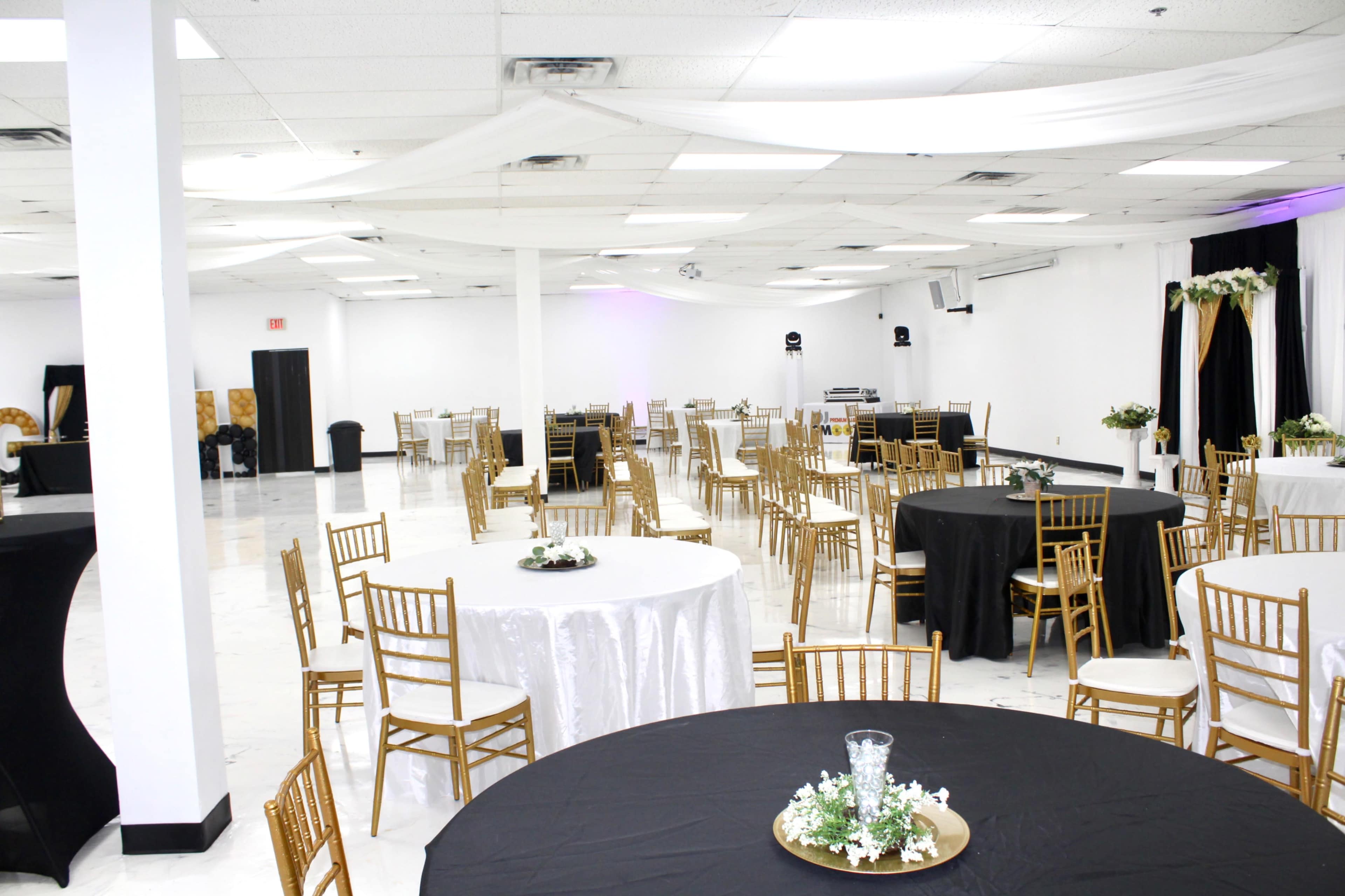A spacious event hall with black and white tablecloths on round tables and gold chairs arranged around the room.