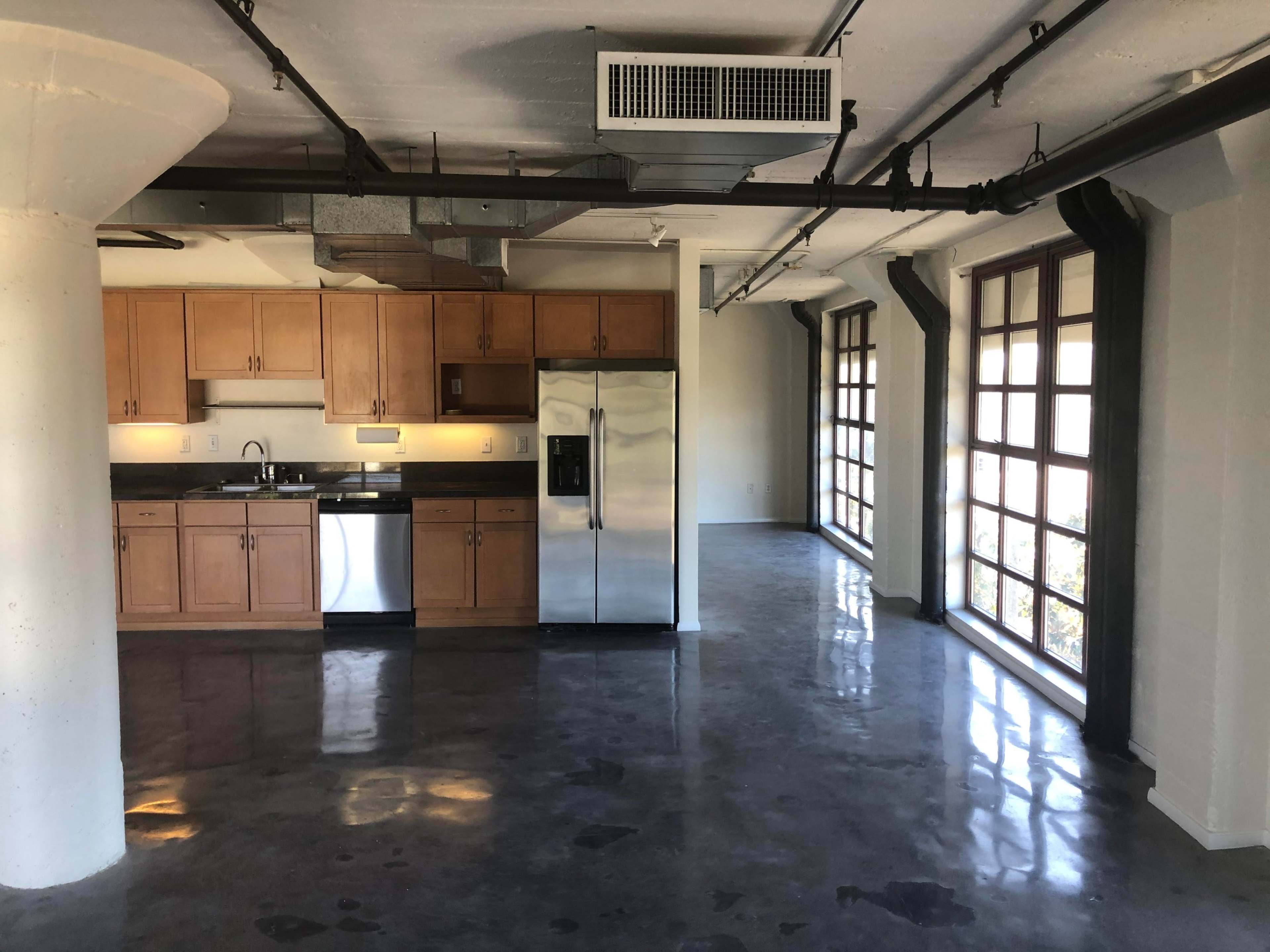 The image shows a modern open-concept living space with a kitchen area featuring wooden cabinets and stainless steel appliances, and large windows allowing natural light to enter.