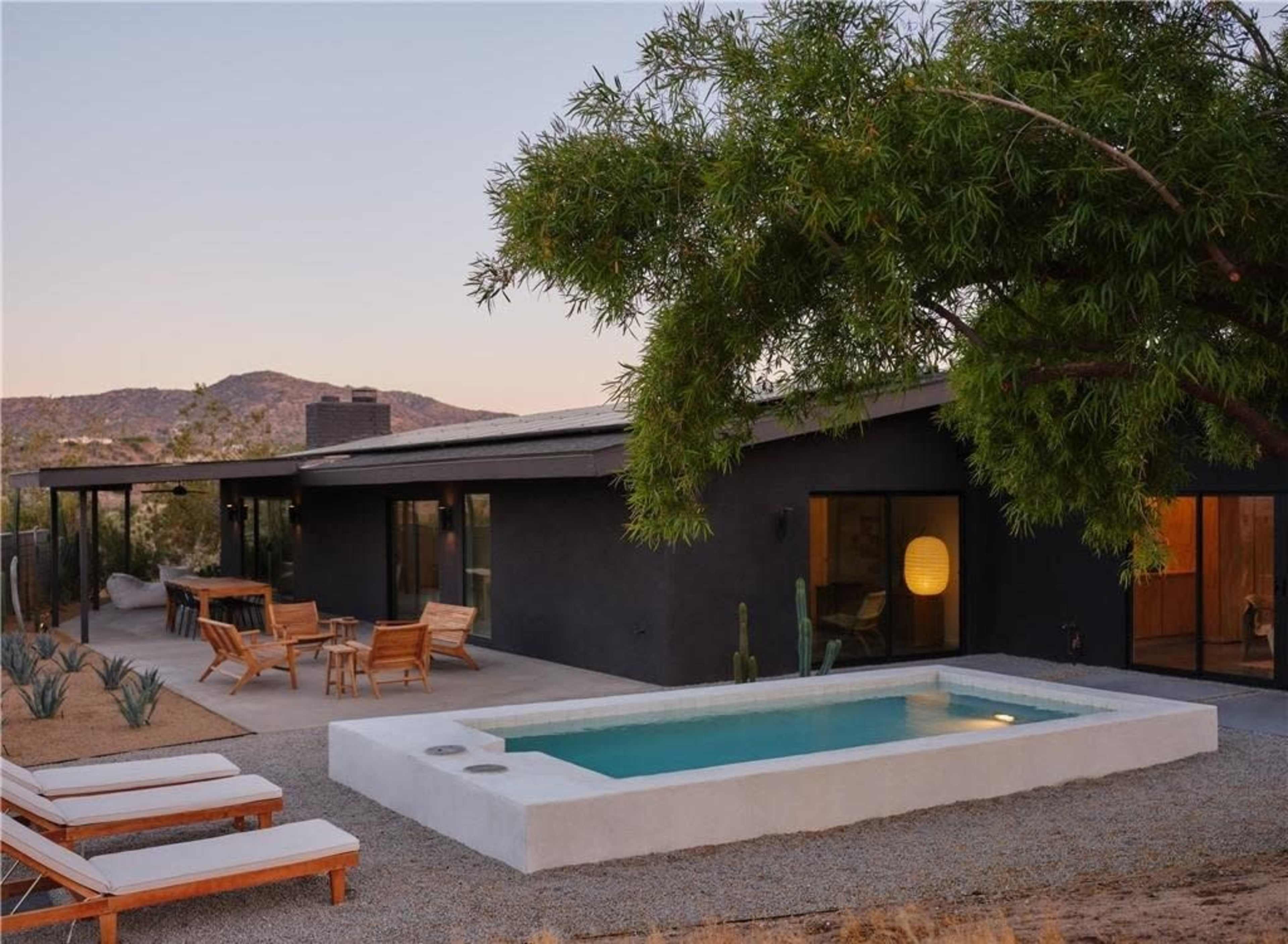 A modern black house with a small pool is surrounded by desert landscaping and mountains in the background.