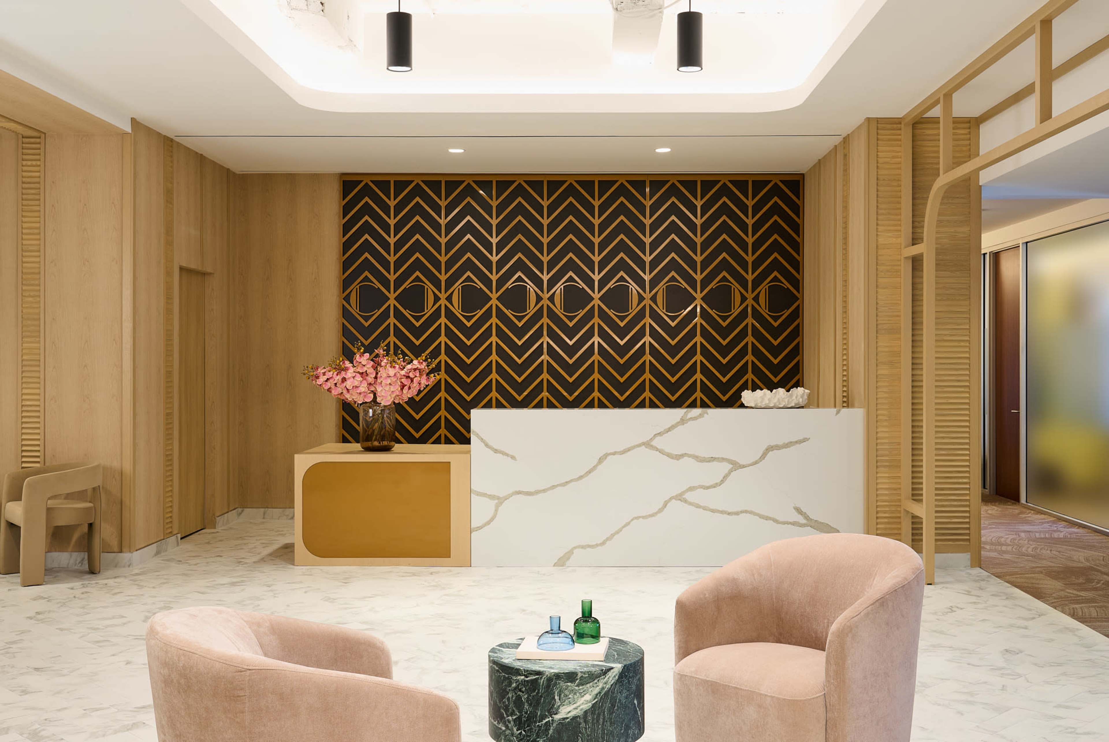 The image shows a modern reception area featuring a marble front desk, a patterned back wall, and plush pink chairs arranged around a small green tabletop.