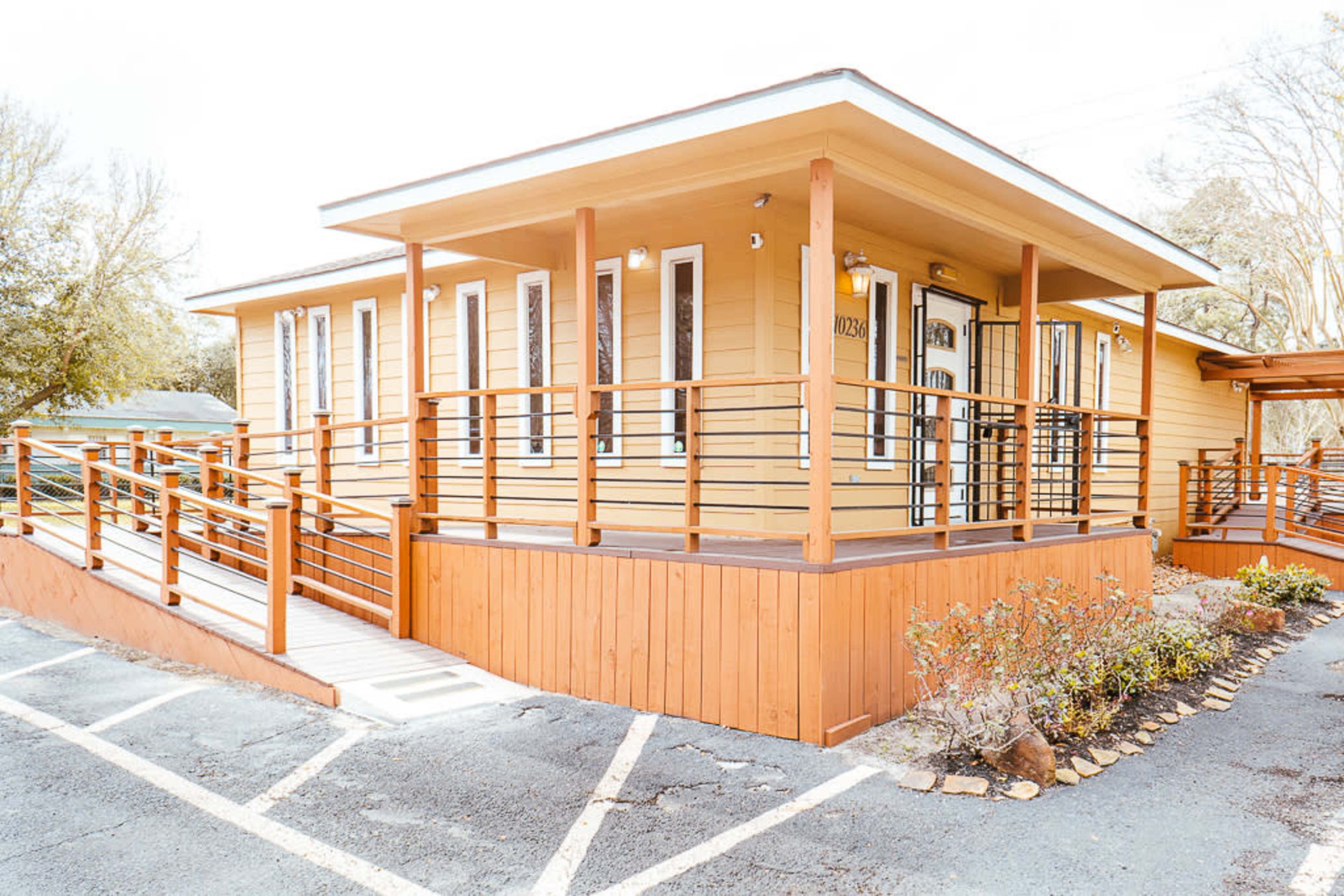 A single-story, wooden building with a ramp and a covered porch, surrounded by a landscaped area and a parking lot.