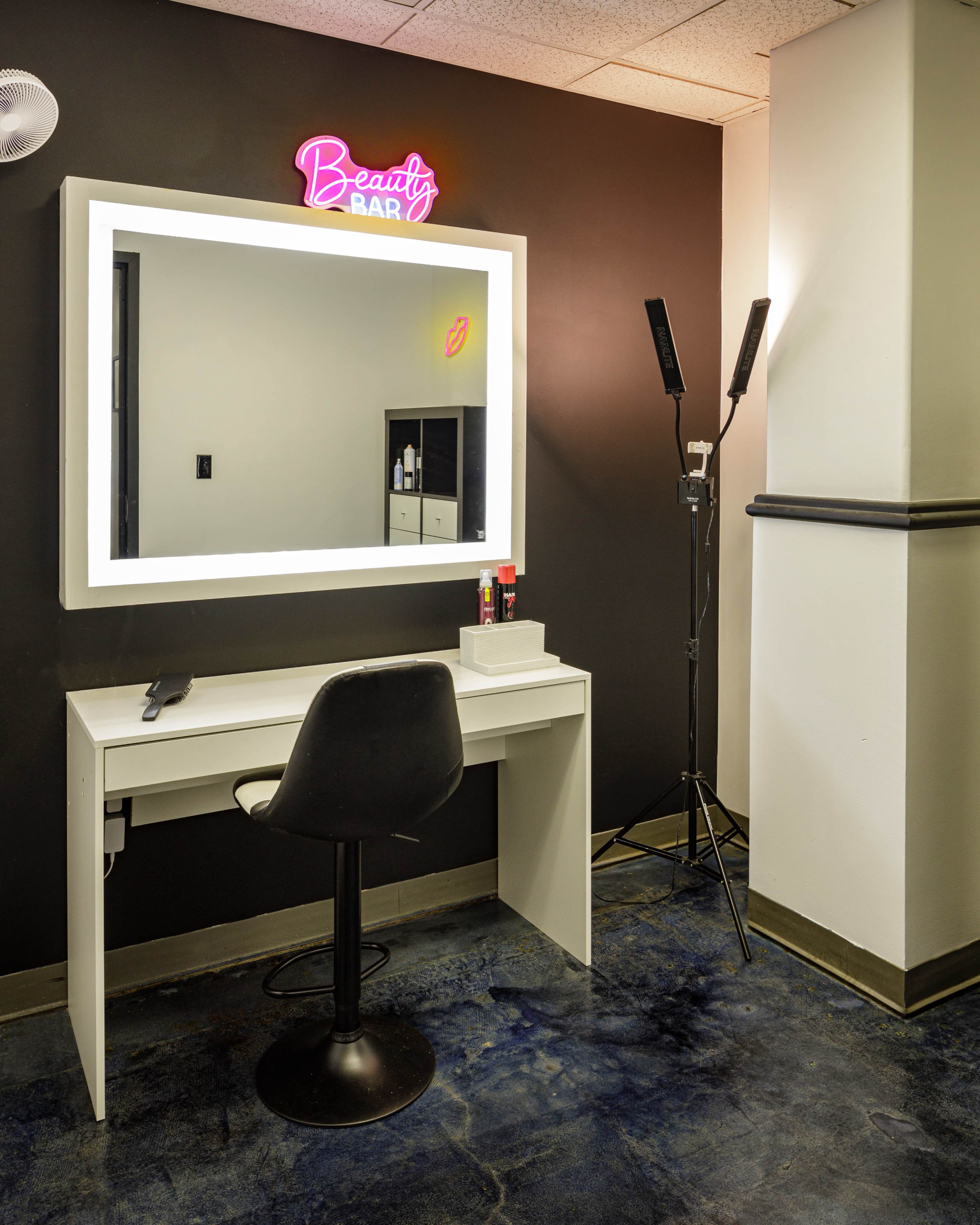 A minimalist beauty bar space featuring a large illuminated mirror, a white vanity table with a black chair, and a neon sign that reads "Beauty Bar."