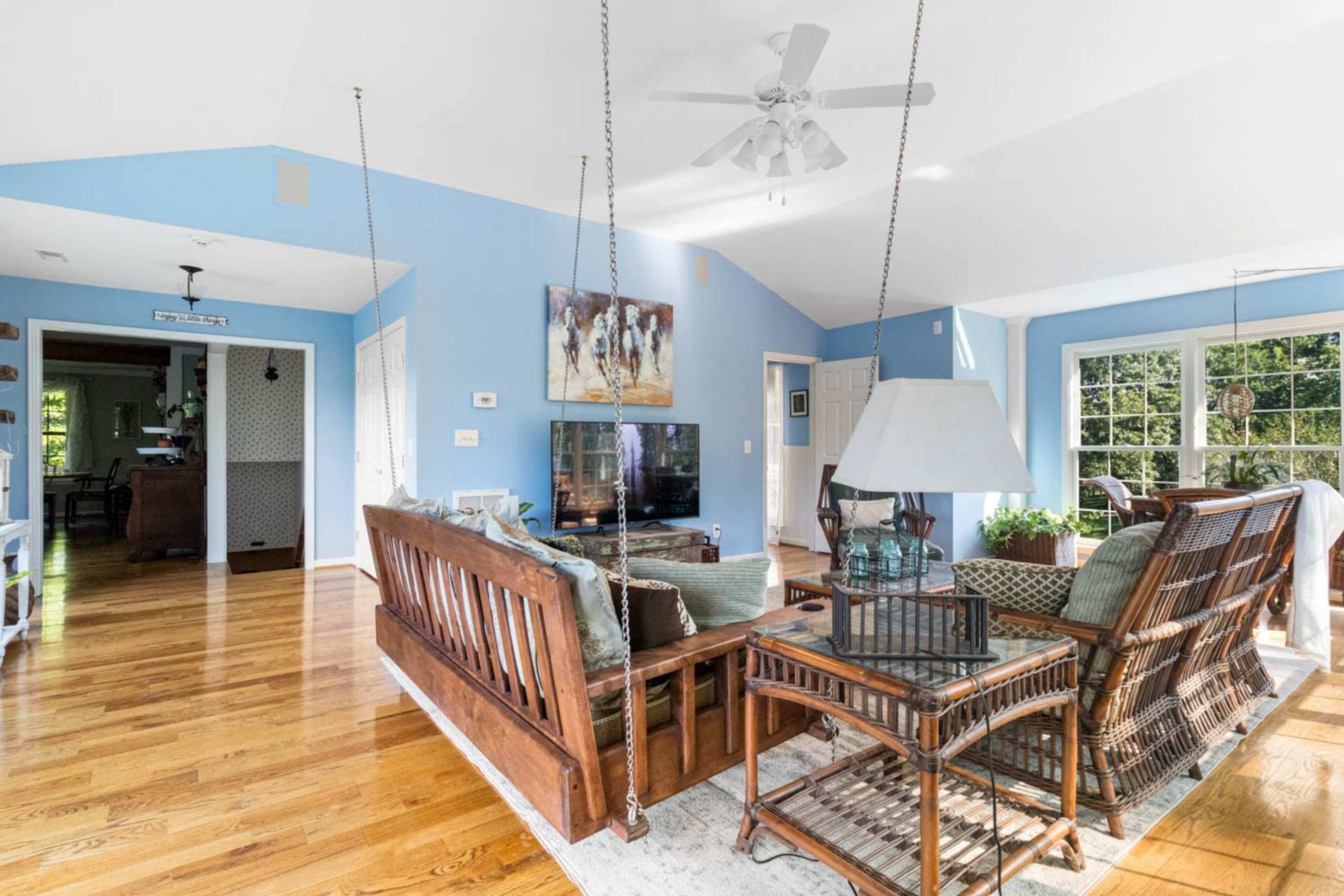 A brightly lit living room with wooden flooring, a blue wall, a ceiling fan, and a seating area featuring a wooden sofa and wicker chairs.