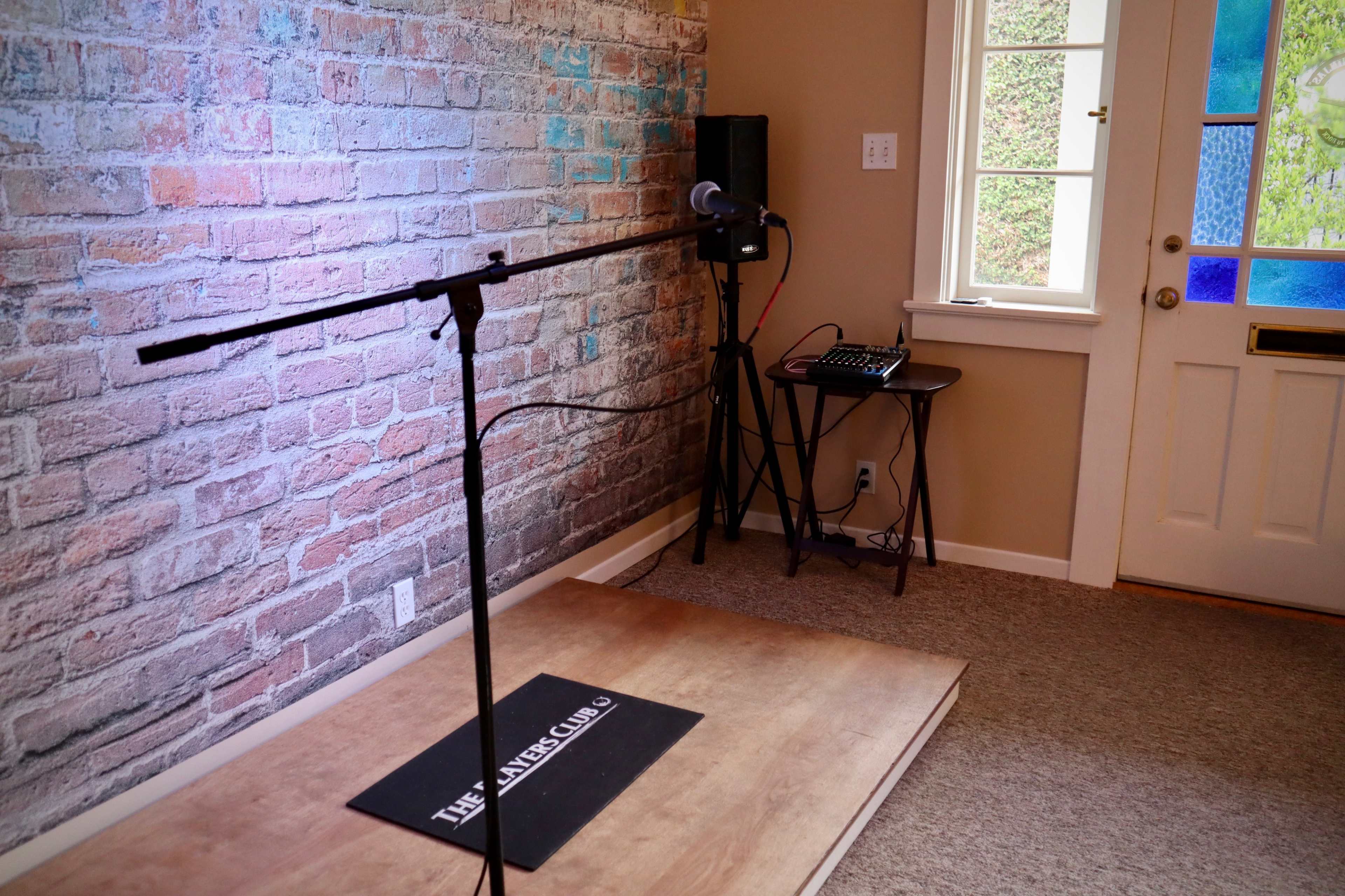 The image shows a small stage with a microphone on a stand in front of a textured brick wall, alongside a sound mixer on a table, near a door with colorful glass panes.