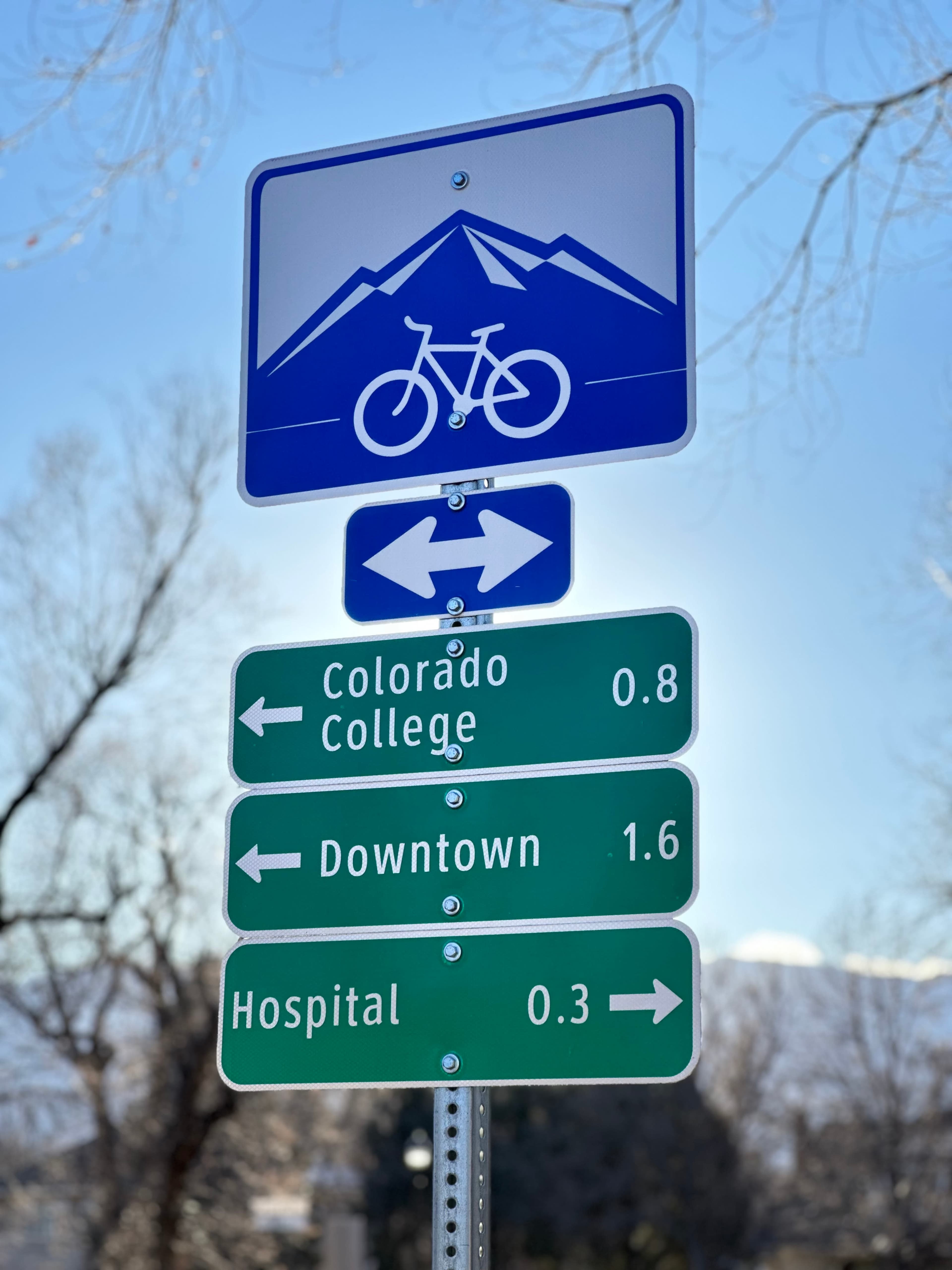 A signpost with directional arrows and distances to Colorado College, Downtown, and a Hospital, accompanied by a bicycle symbol above a mountain graphic.