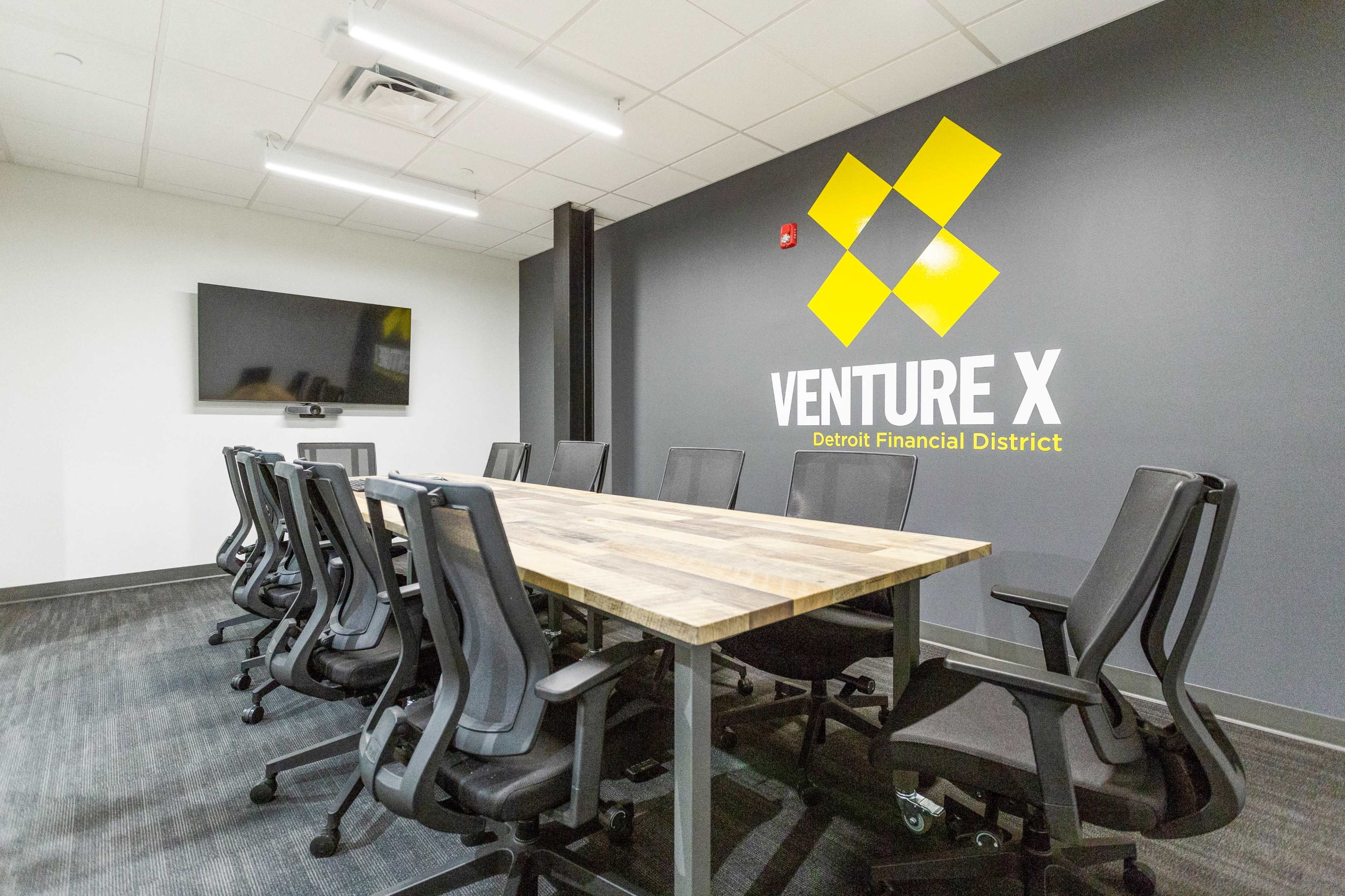 A modern conference room featuring a large wooden table, ergonomic chairs, and a wall with the logo of "Venture X" in the Detroit Financial District.