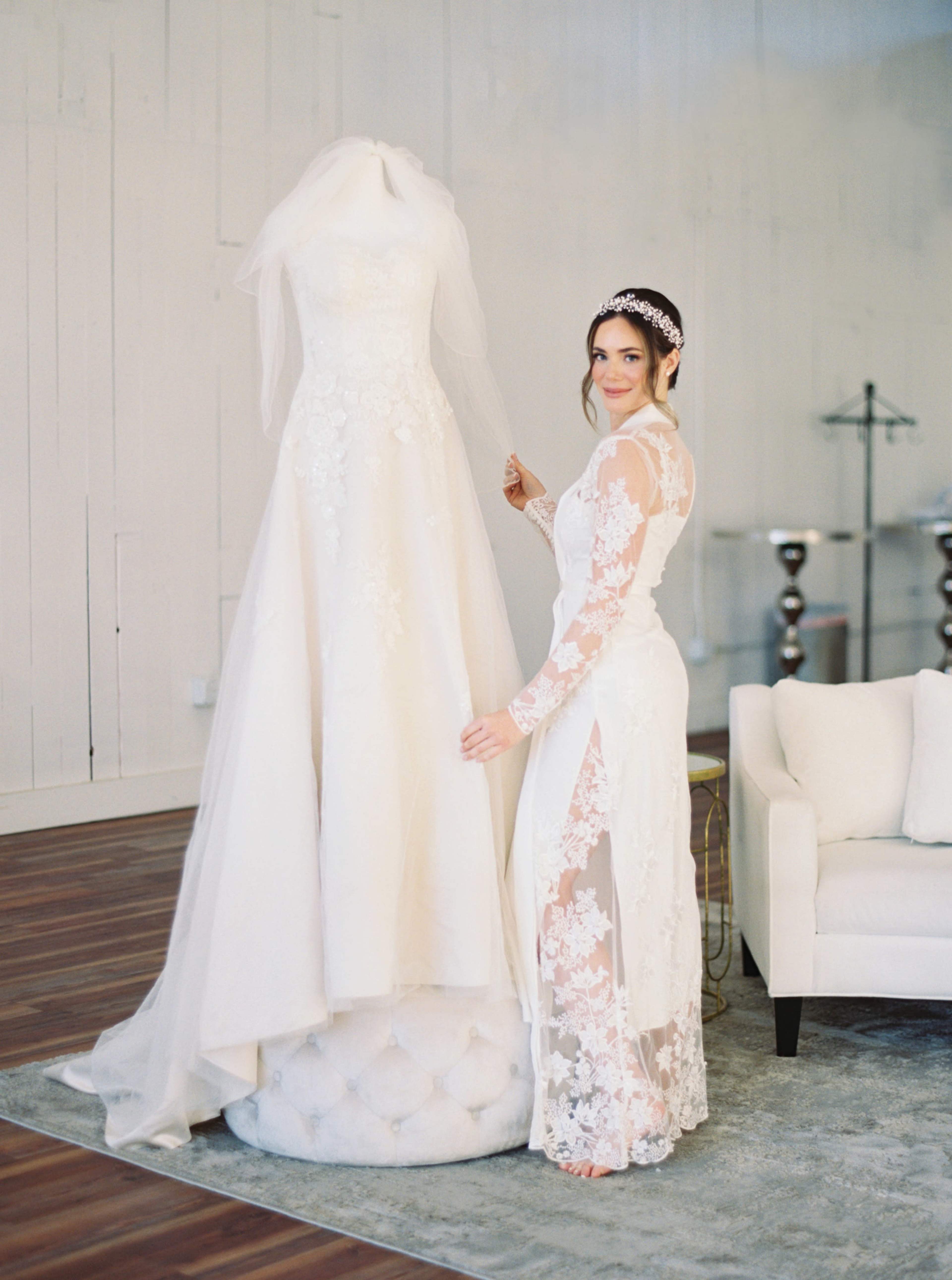 A bride in a lacy wedding dress stands next to a mannequin displaying a similar gown in a stylish indoor setting.