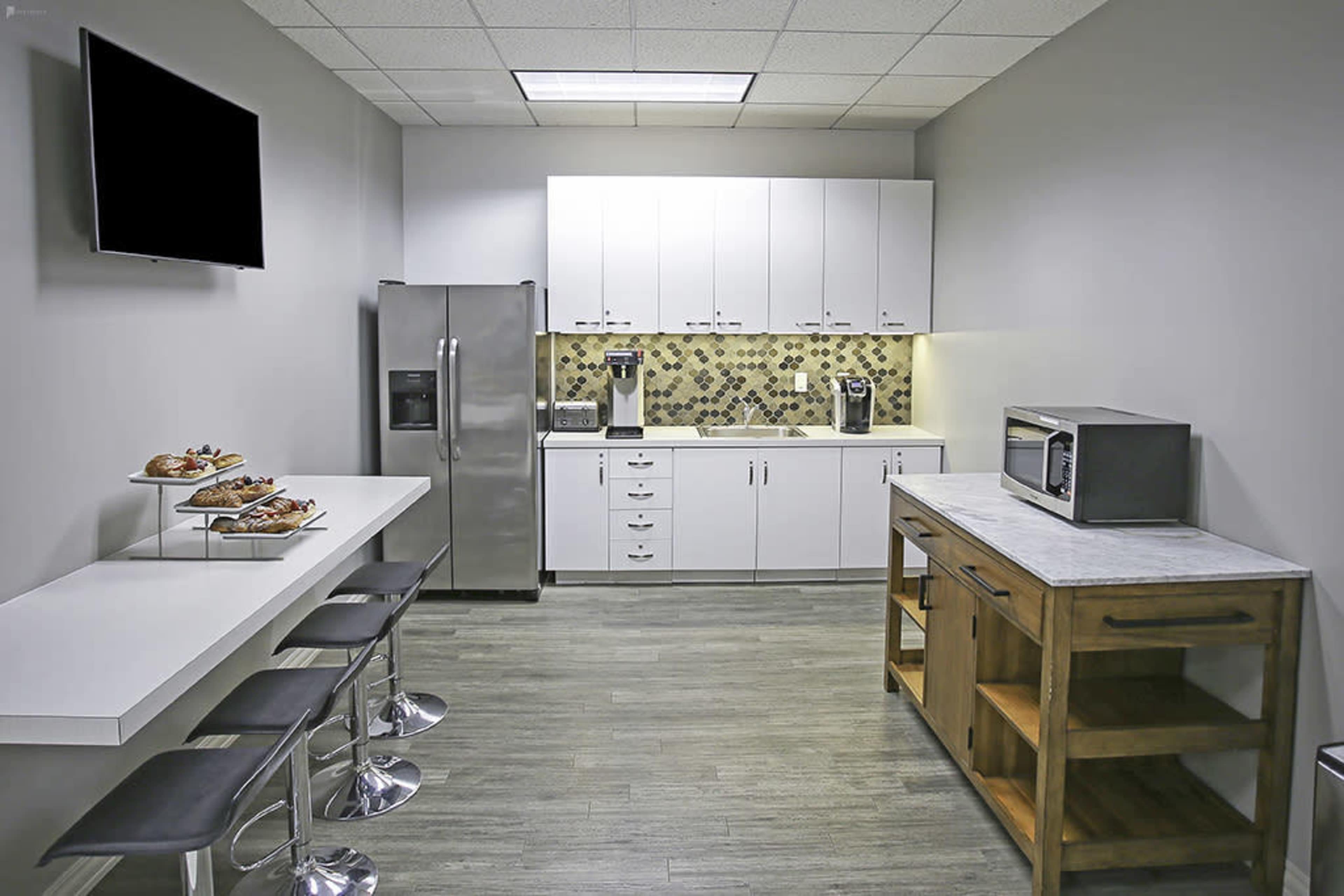 A modern kitchenette features a stainless steel refrigerator, white cabinets, a countertop with a coffee maker, and a snack bar with stools.