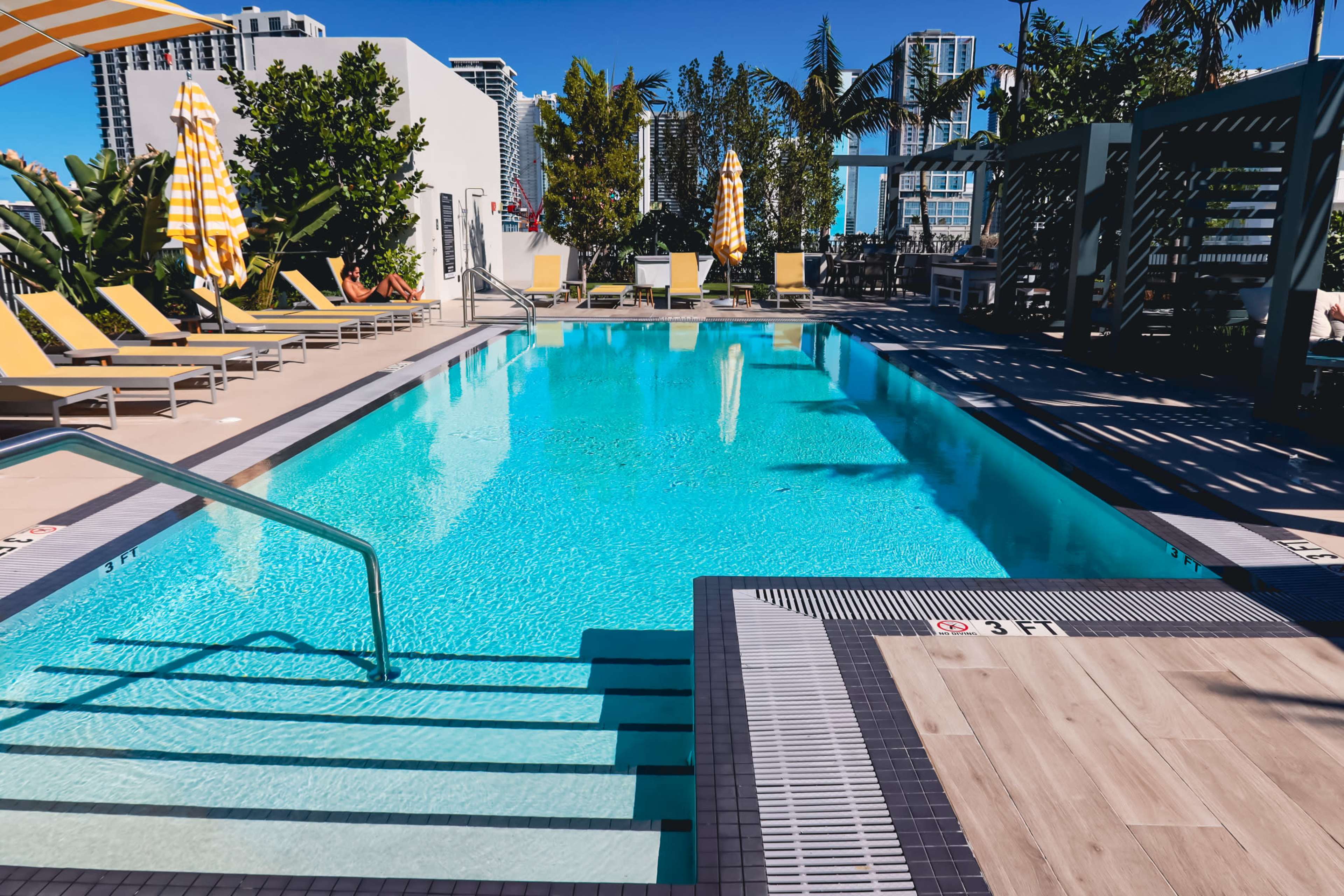 The image shows a clear swimming pool surrounded by lounge chairs and palm trees, with bright yellow umbrellas providing shade.