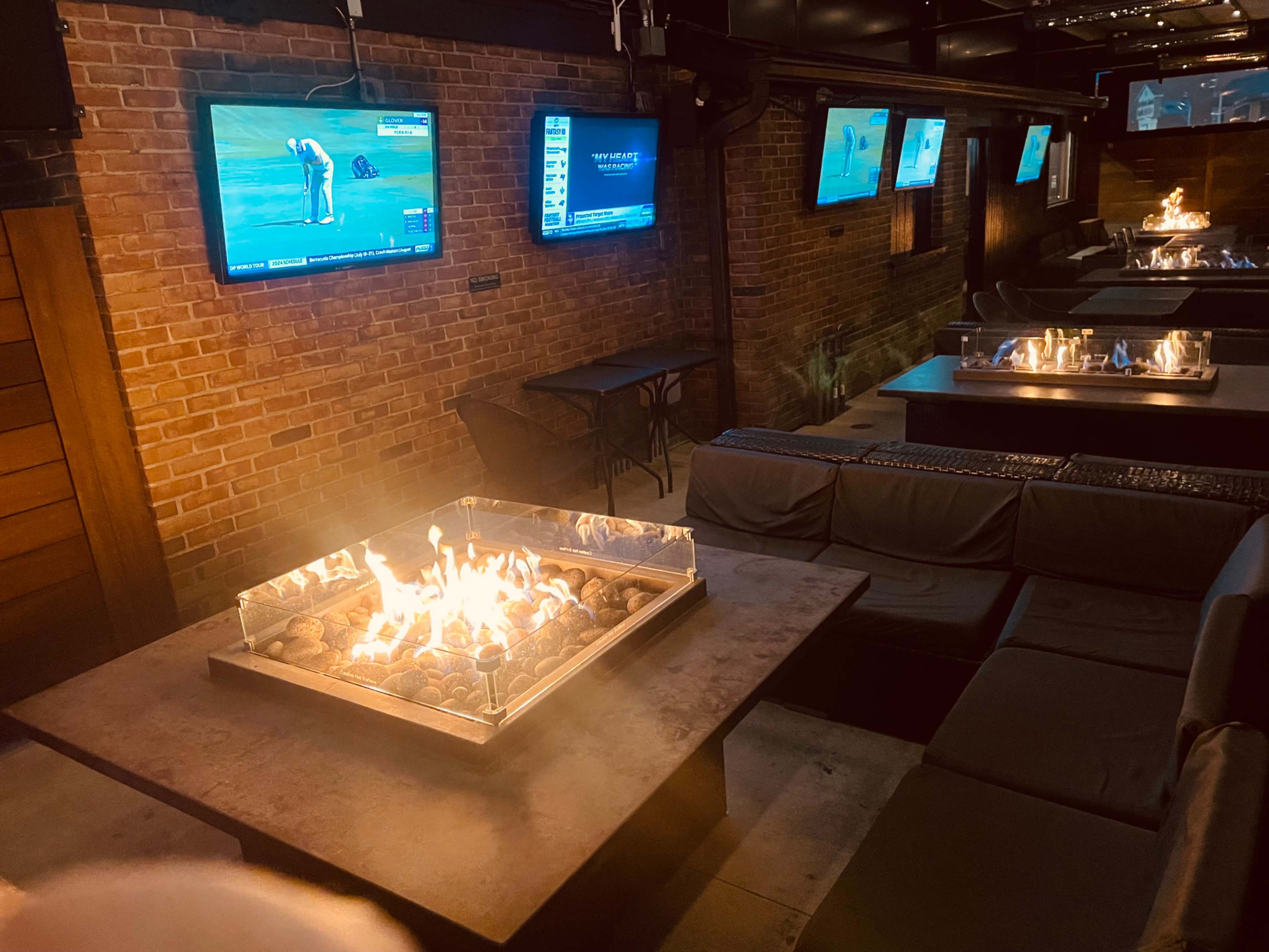 A modern indoor lounge area with brick walls, a central fire table, and multiple screens displaying sports broadcasts.