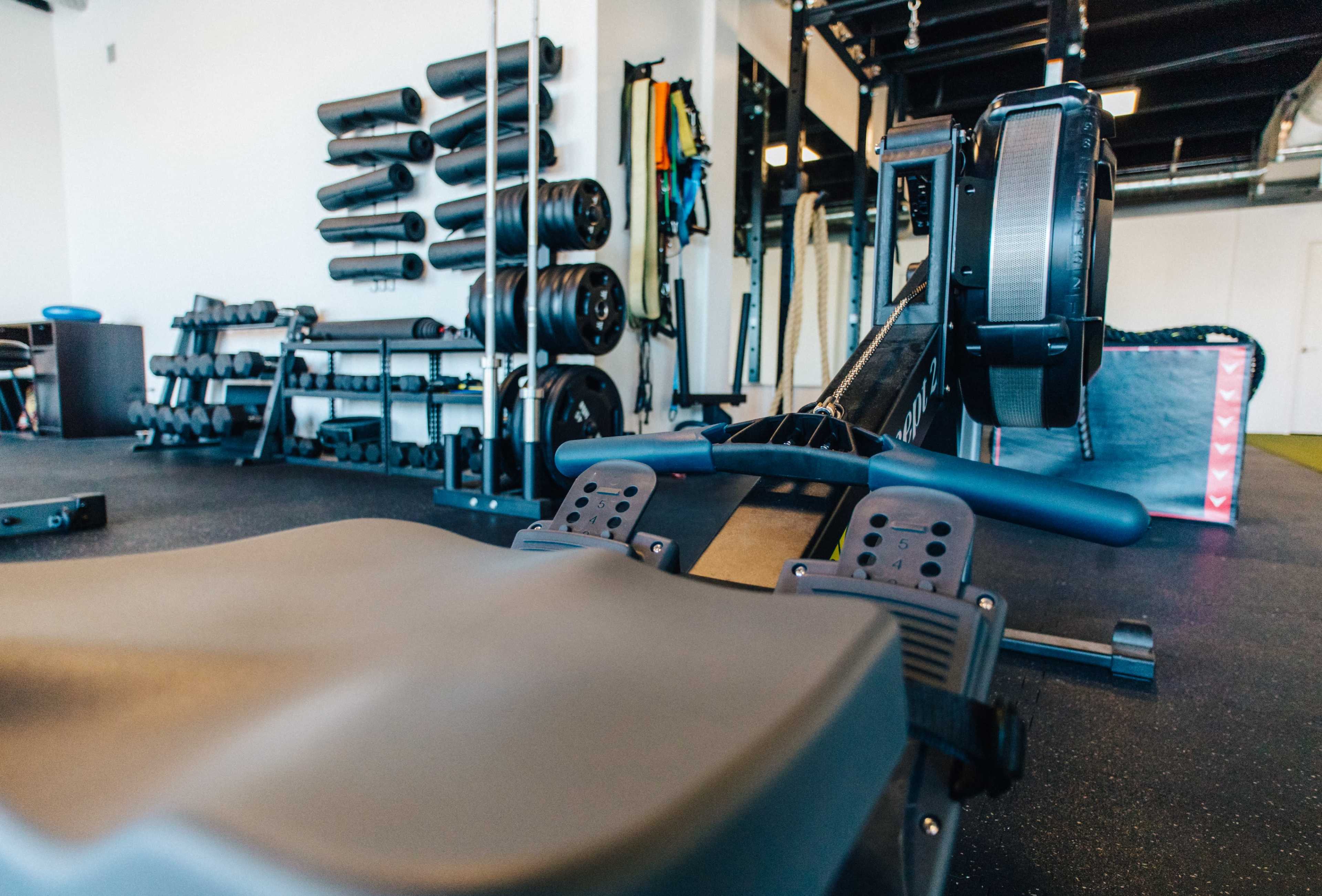 A rowing machine in a gym, with free weights and exercise equipment arranged on the walls in the background.