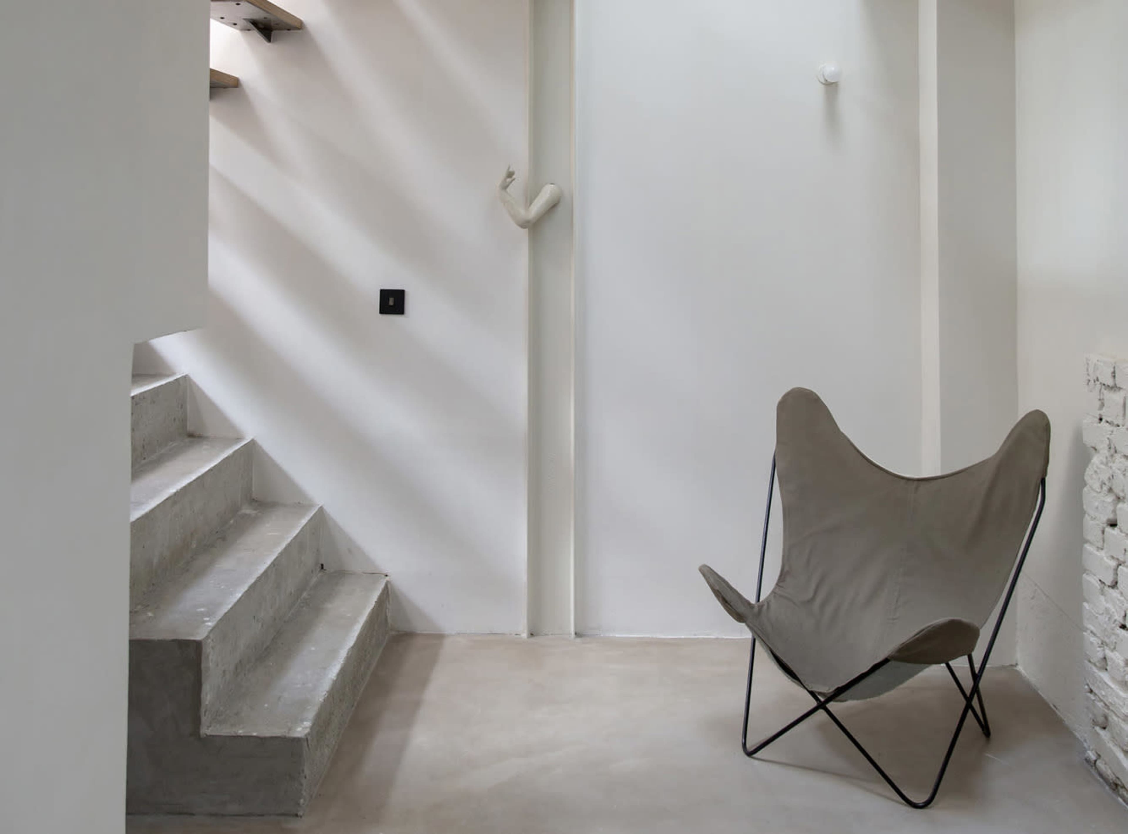 The image shows a minimalist interior space featuring a single butterfly chair beside a staircase with concrete steps.