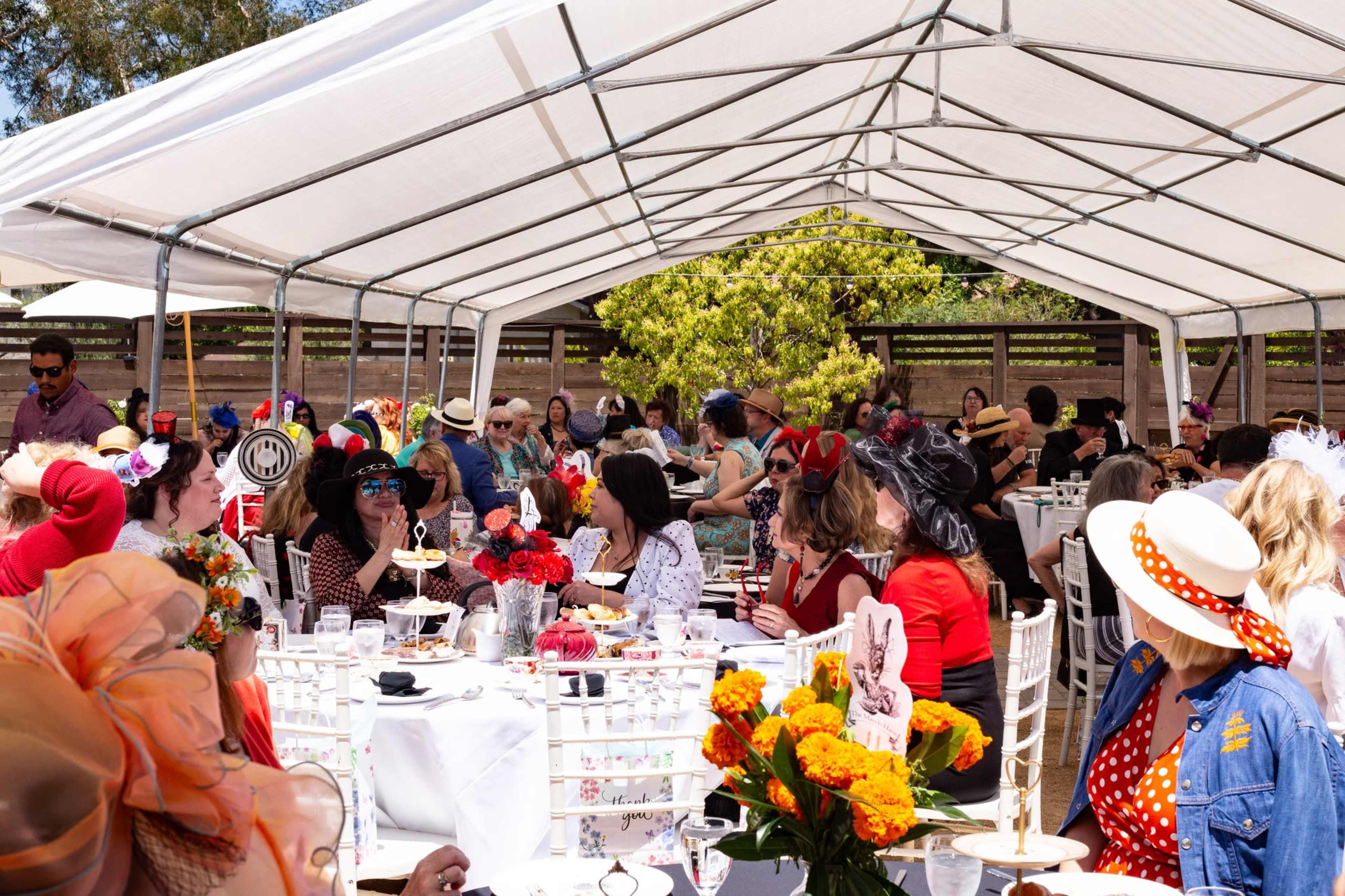 A large outdoor gathering under a tent features numerous guests wearing hats, seated at elegantly set tables adorned with flowers and food.