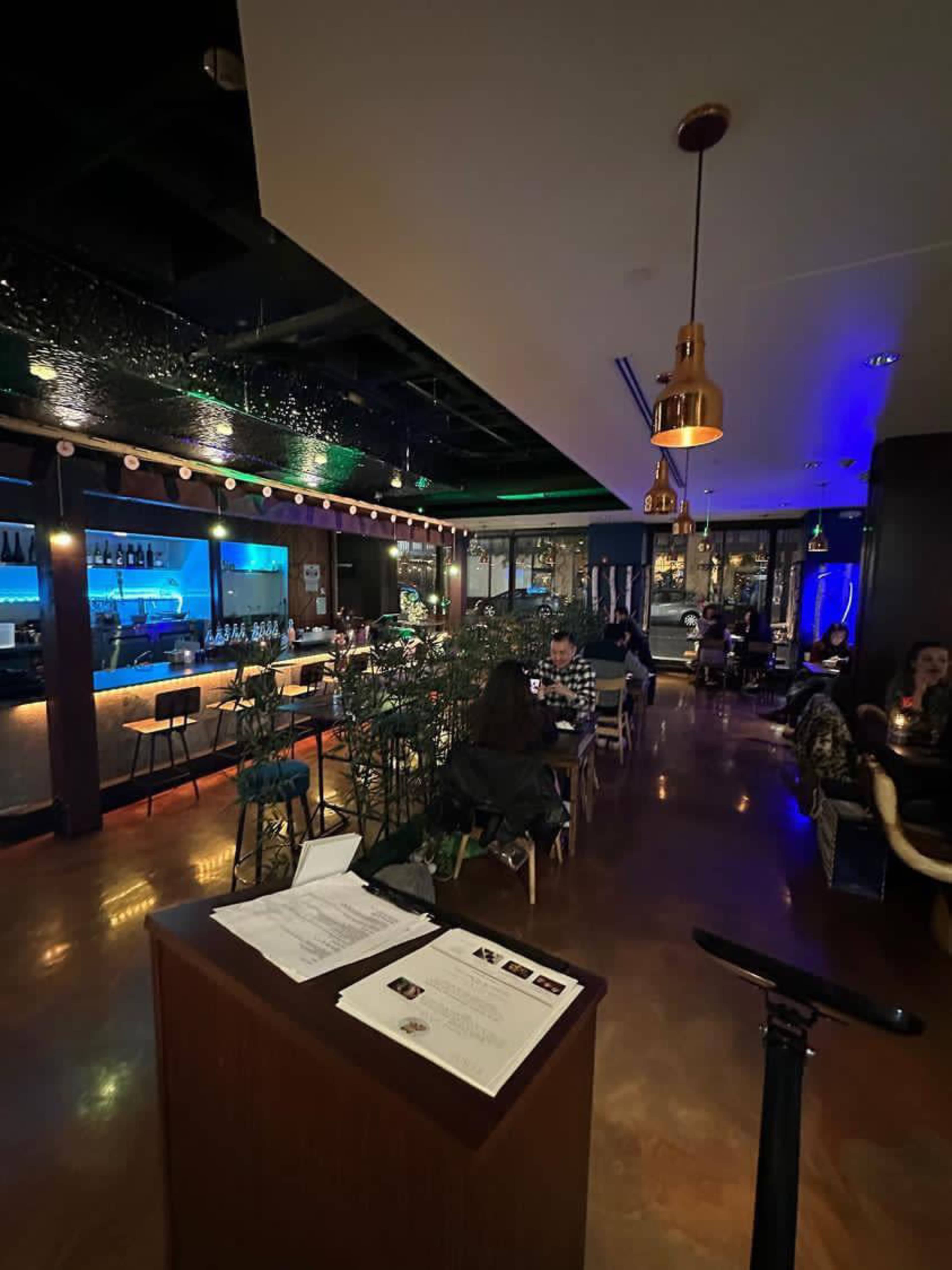 The interior of a restaurant featuring a bar area with blue lighting, seated patrons, and decor that includes hanging pendant lights and greenery.