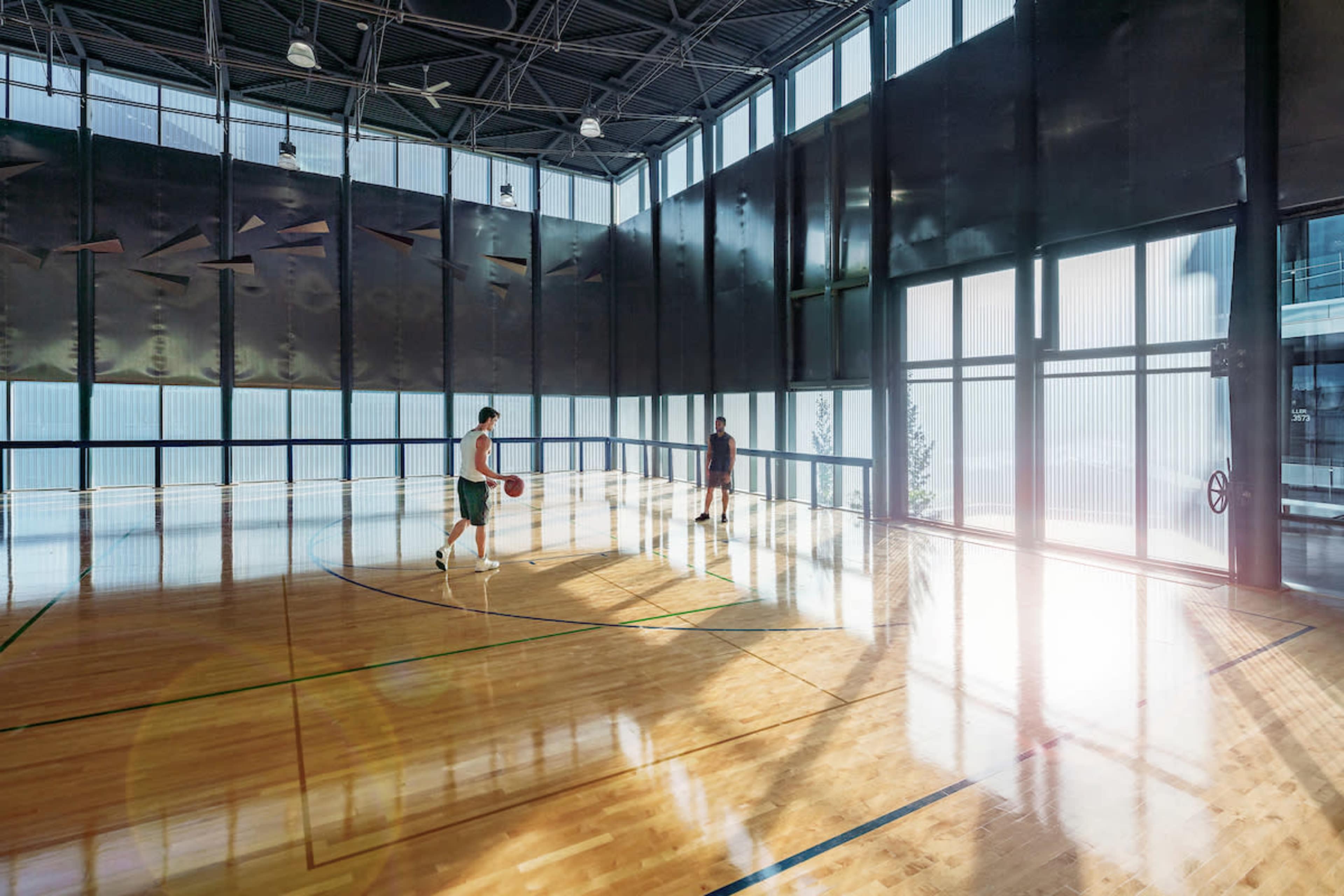 Two individuals play basketball in a brightly lit indoor court with large windows.