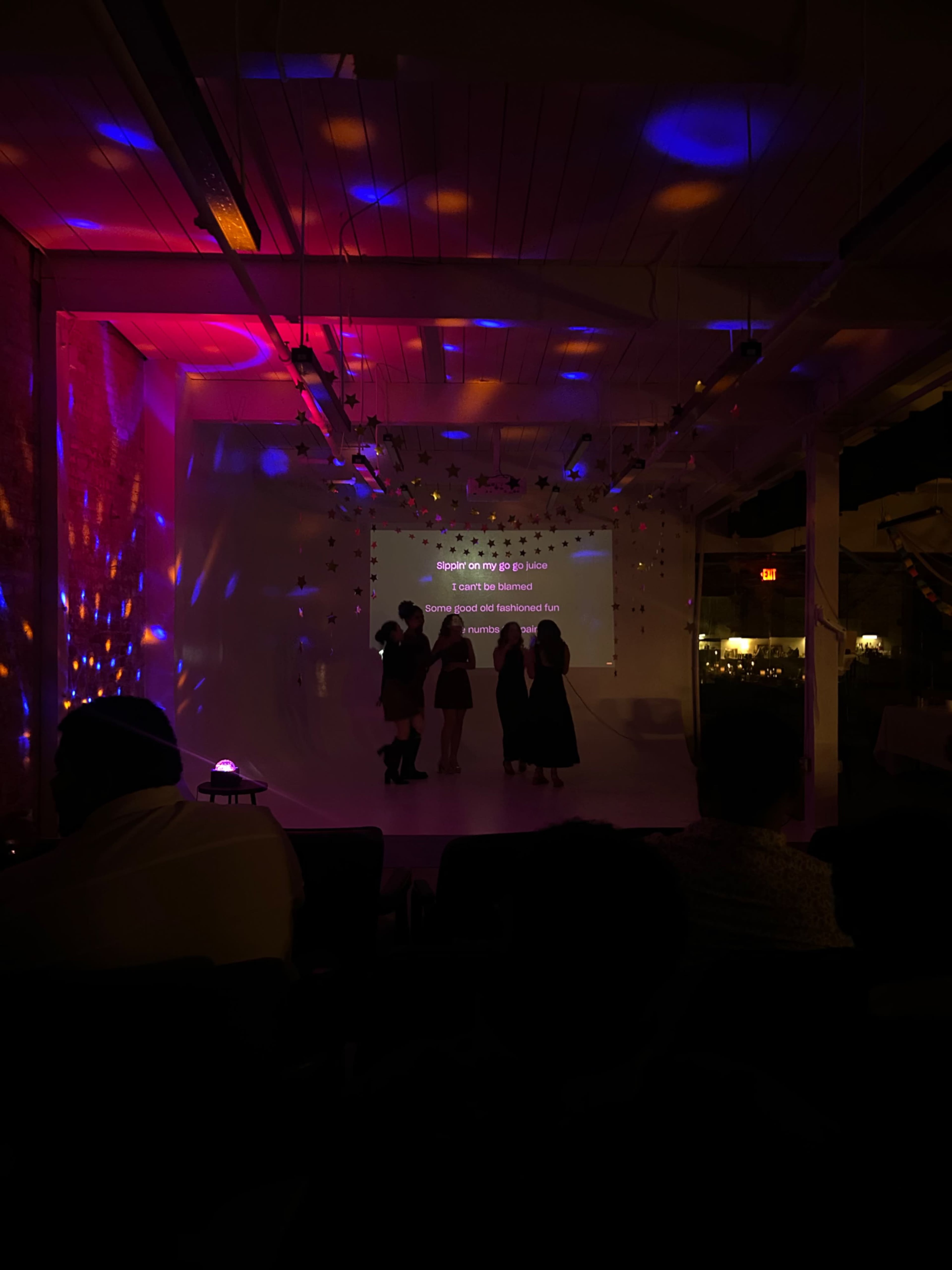 A group of people stands in front of a screen displaying text, while colorful lights illuminate the walls of a dimly lit room.