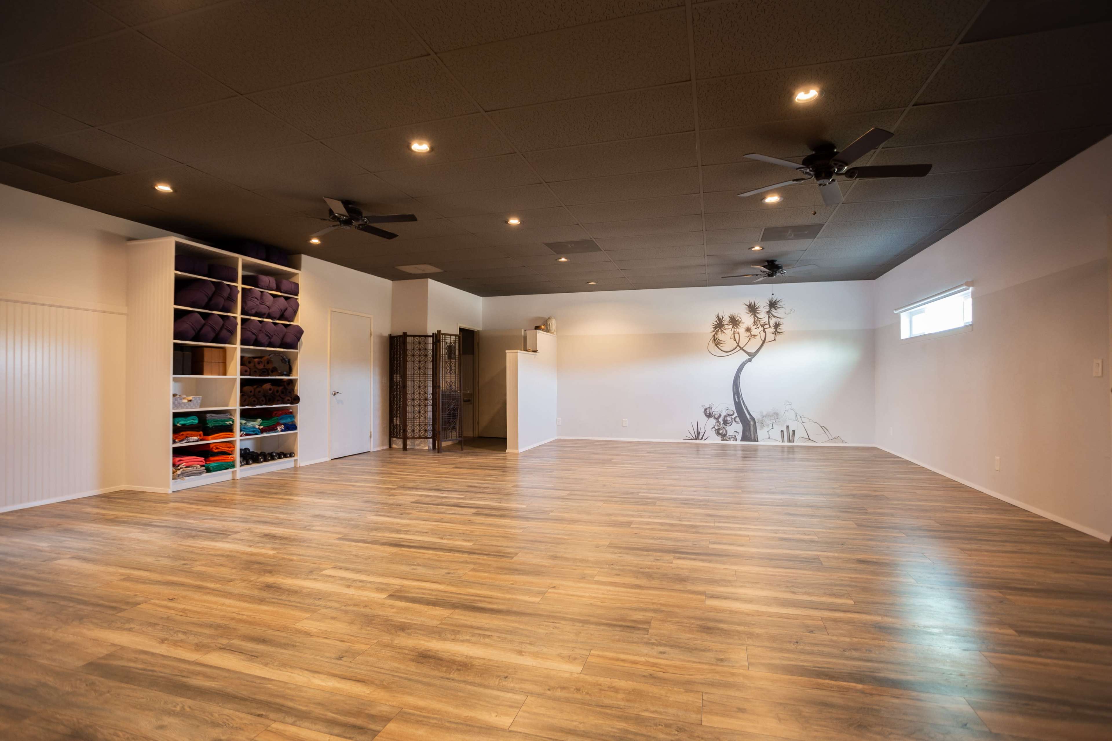 A spacious yoga studio featuring wooden floors, a wall mural of a tree, shelving with neatly stacked yoga mats, and two ceiling fans.