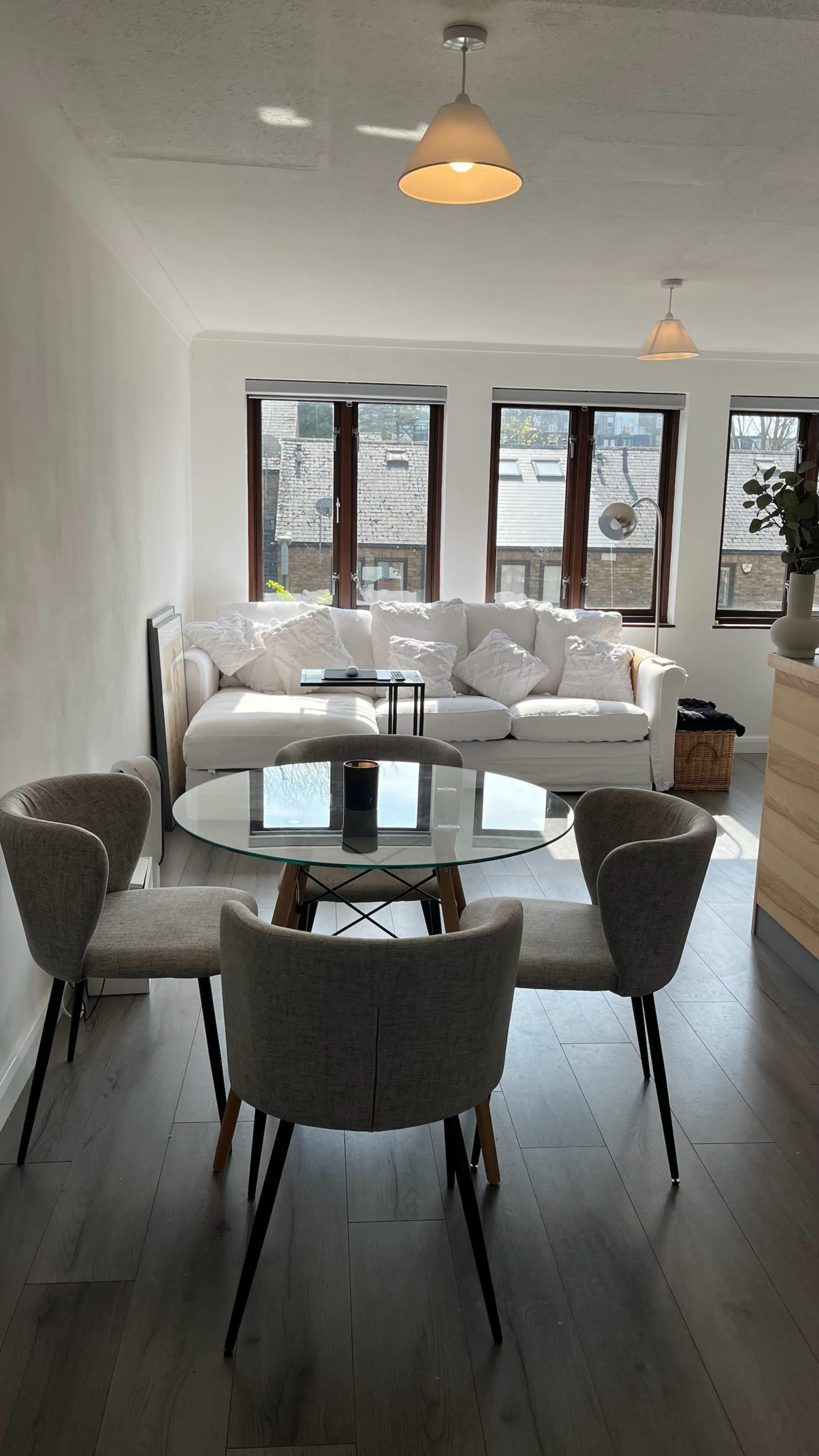 A modern dining area features a glass round table surrounded by four gray chairs, with a cozy white sofa and large windows in the background.