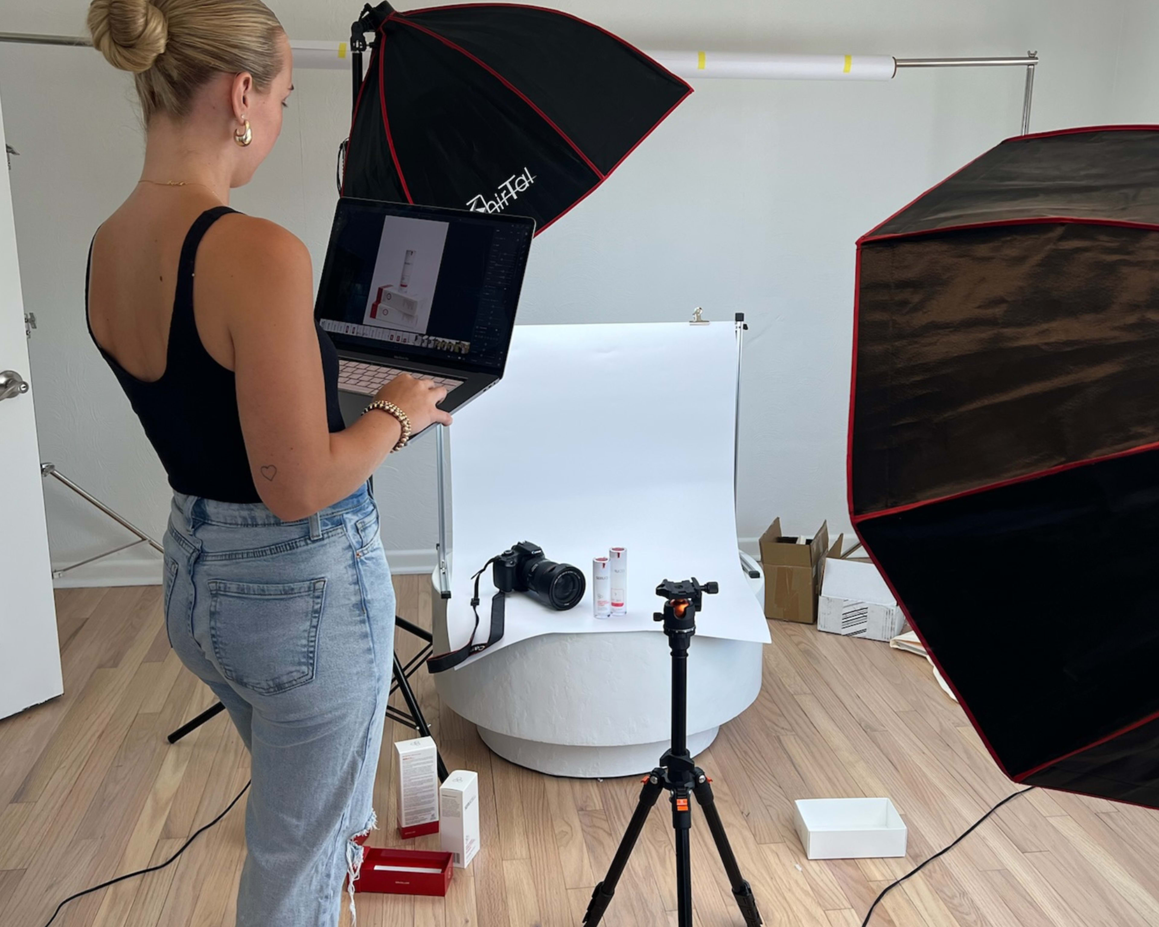 A person stands holding a laptop while setting up a product photography scene with a camera and lighting equipment.